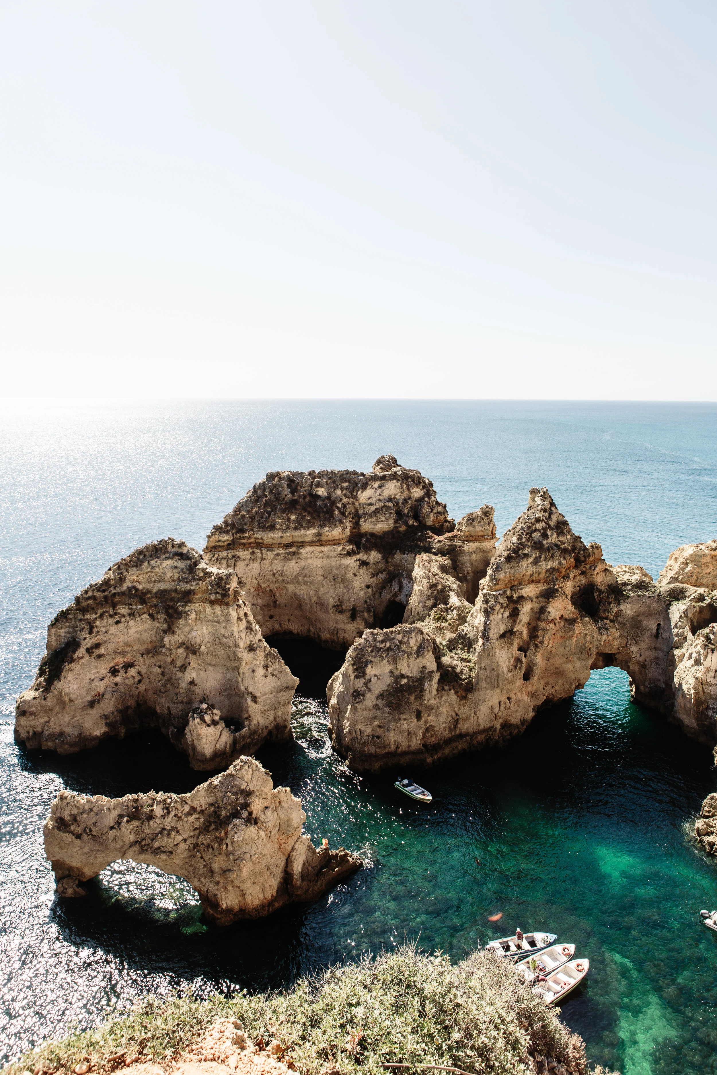 Cliffs with natural arches and caves overlooking the ocean, with boats anchored near the rocks and a clear, calm sea.