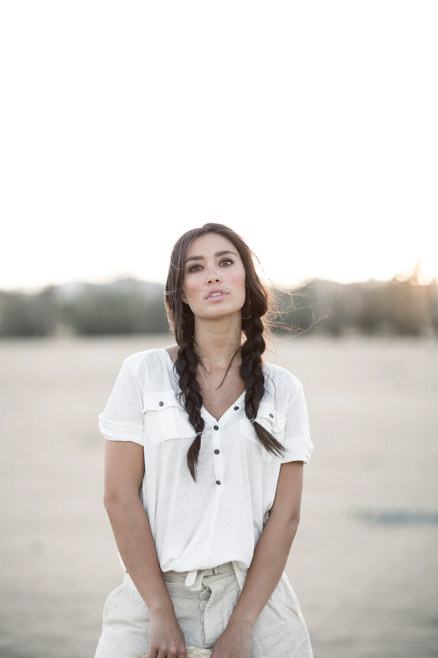 A young woman with dark hair in braids standing outdoors in a wide open field at sunset, wearing a white shirt and beige pants.