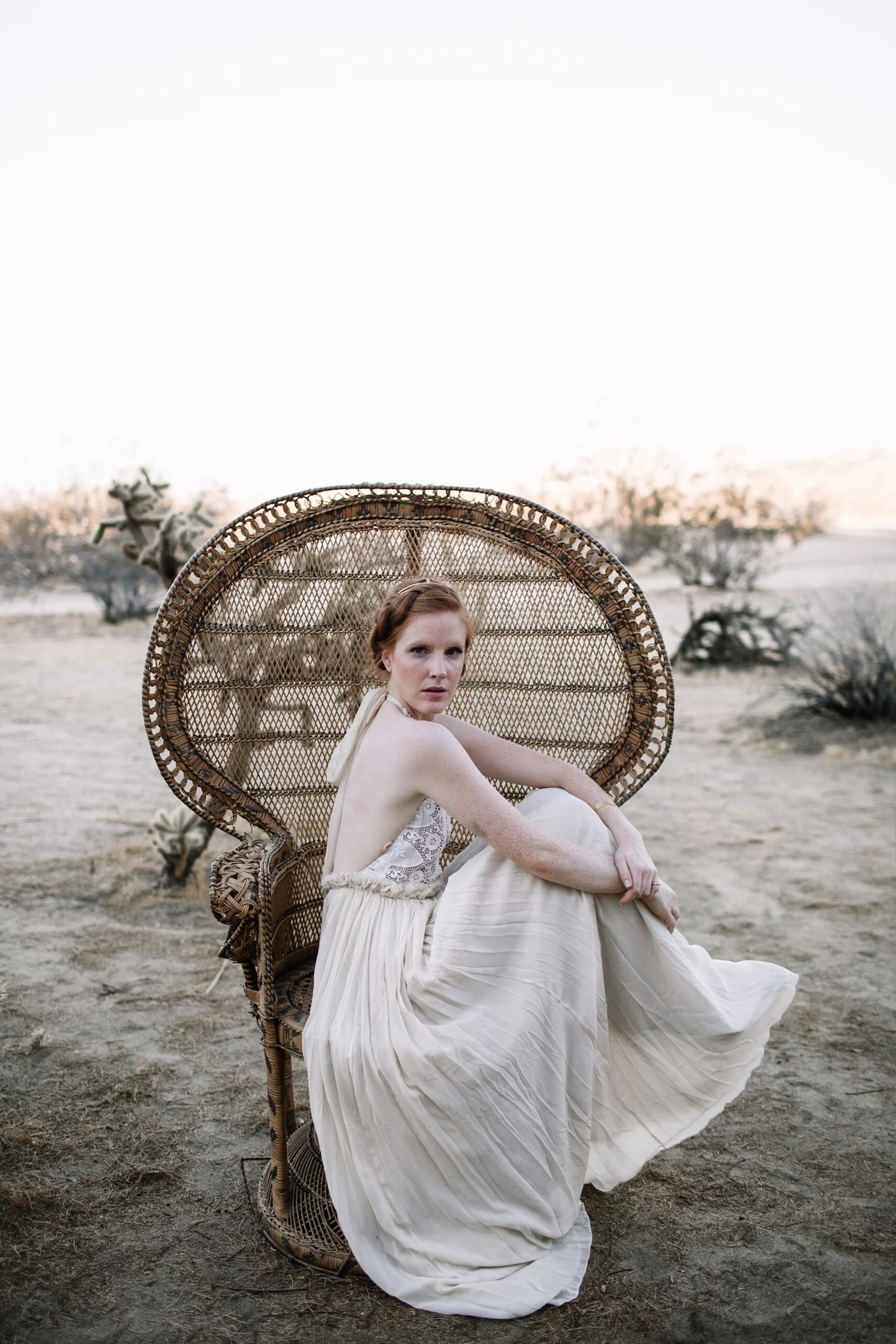 A woman with red hair wearing a white dress sitting on a vintage wicker peacock chair in a desert landscape during sunset.