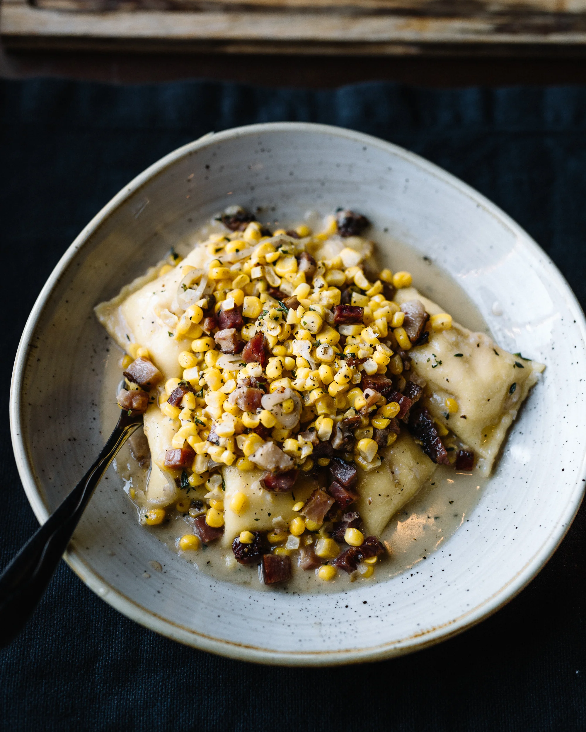 A bowl of ravioli with a topping of corn and diced bacon, served in a creamy sauce.