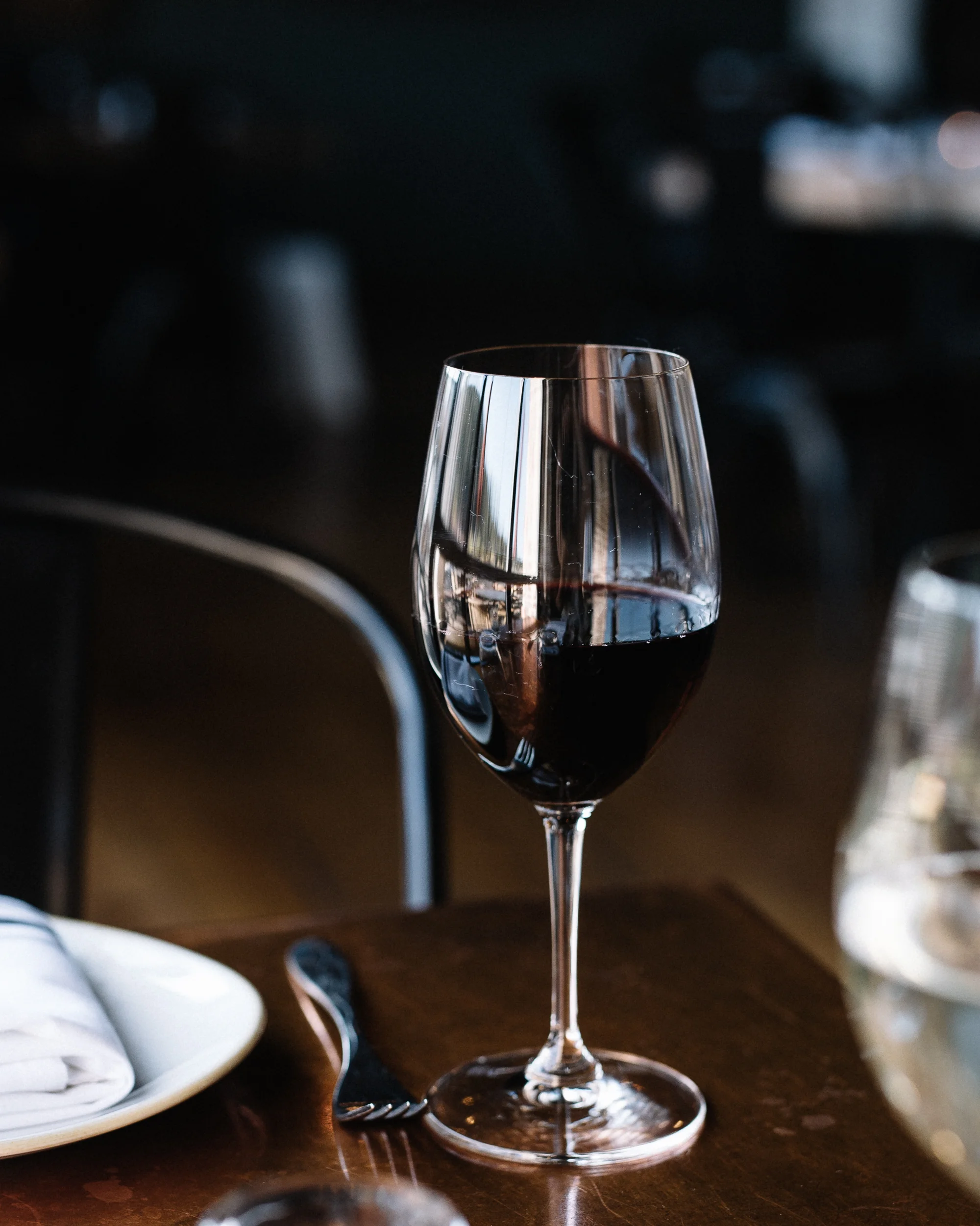 A glass of red wine on a wooden table with a fork and a plate with a white cloth napkin nearby.