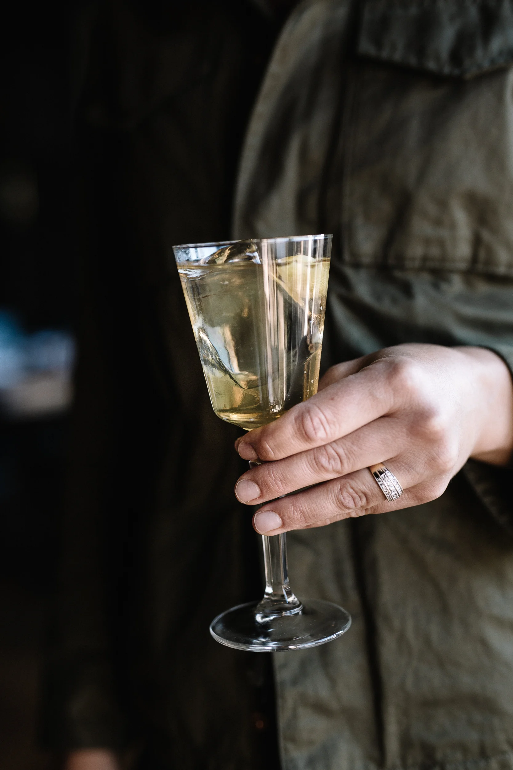 A person holding a wine glass with a clear beverage, possibly white wine or champagne, with a lemon wedge inside. The person is wearing a ring and a green jacket.