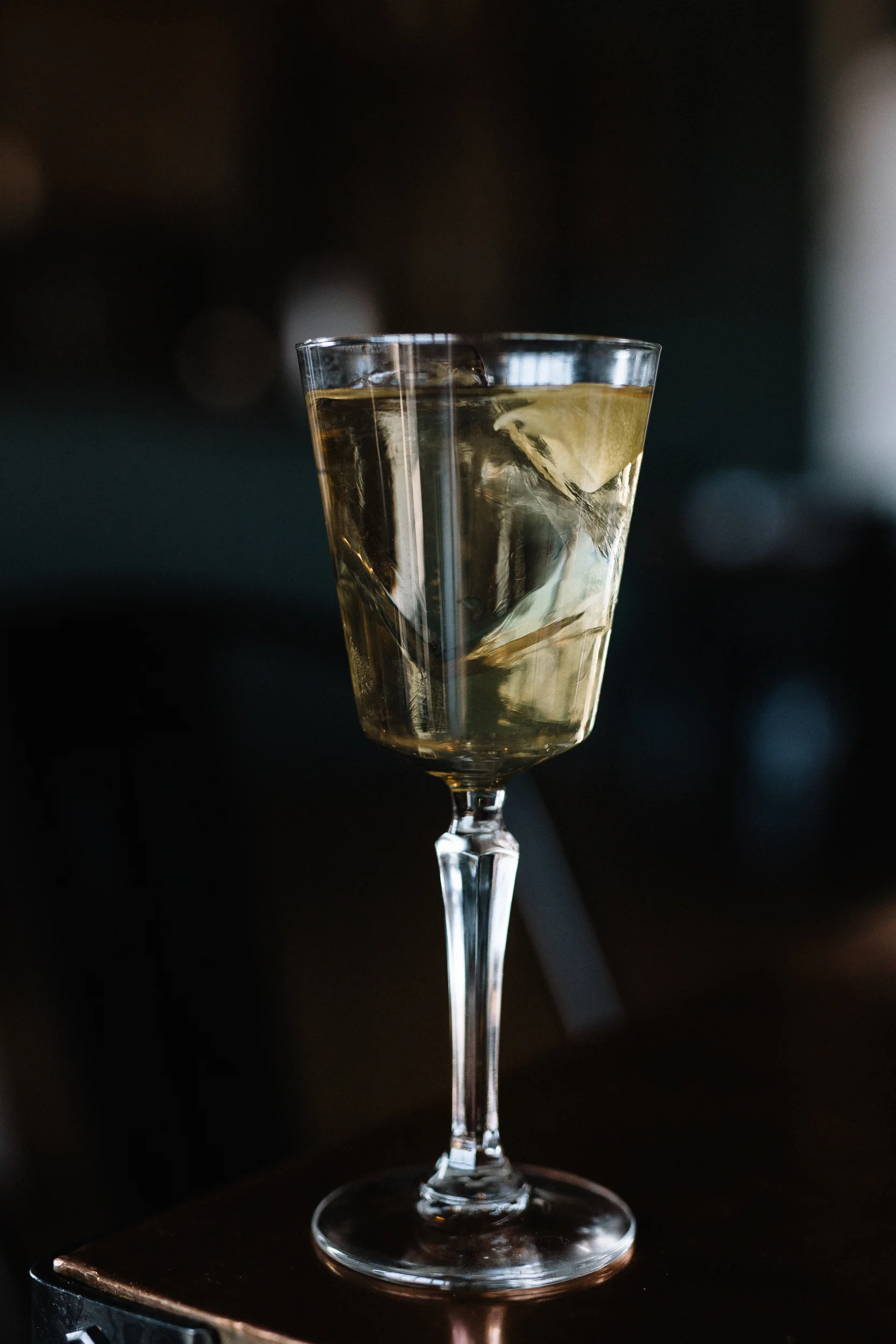 A tall, elegant glass filled with a clear beverage and ice cubes, set against a dark, blurred background.