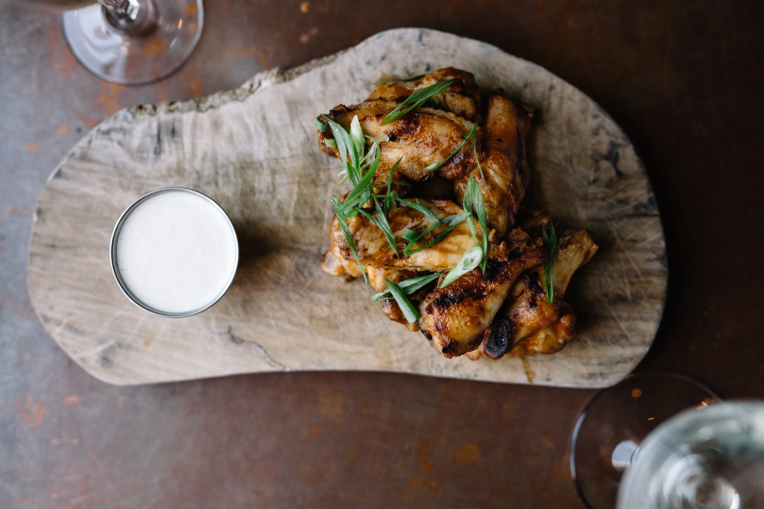 Grilled chicken wings garnished with green herbs served on a rustic wooden platter with a small glass of white dipping sauce, on a dark wooden table.