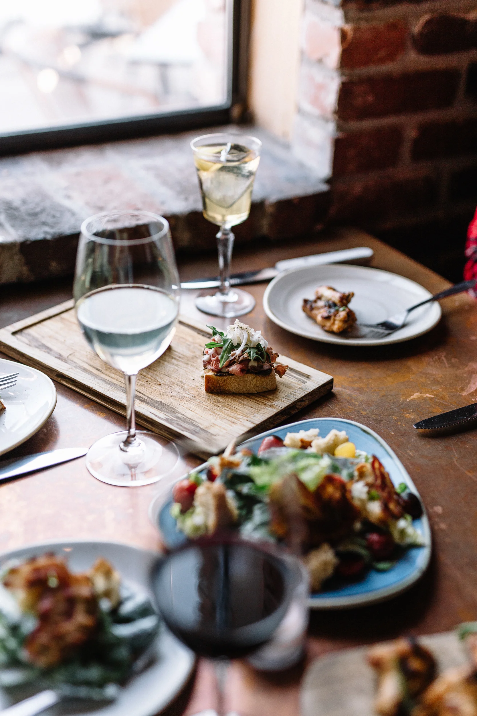 A rustic wooden table set with various plates of food and glasses of water and white wine near a brick wall and window.
