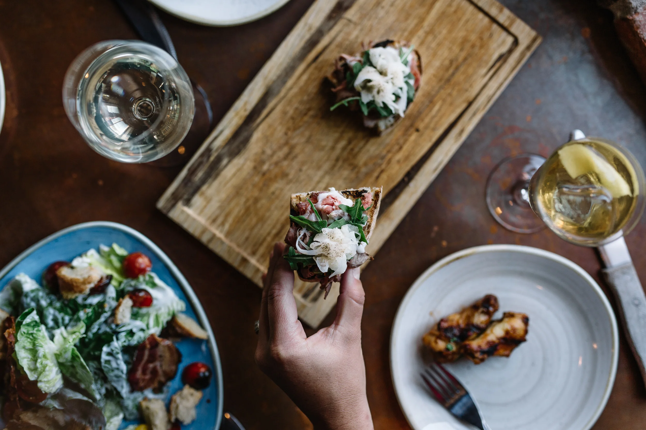 A hand holding a small piece of toasted bread topped with vegetables, herbs, and cheese at a table with various dishes and drinks, including a salad, chicken wing, wine, and a glass of white wine.