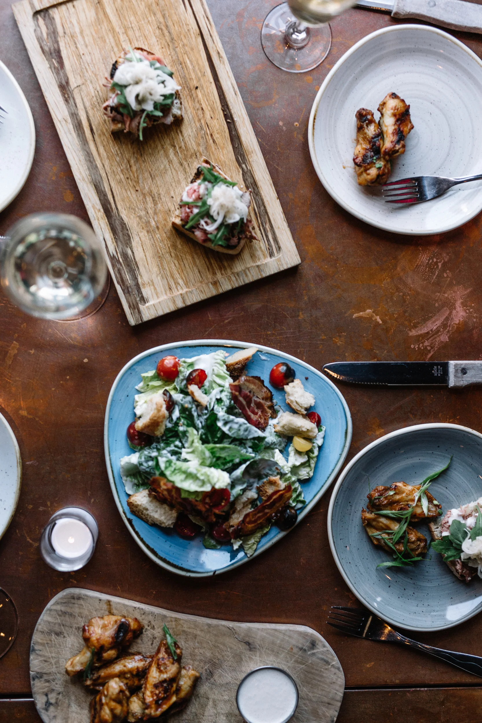 A top-down view of a dining table with plates of salad, grilled chicken wings, and appetizer bites, along with wine glasses and a candle.