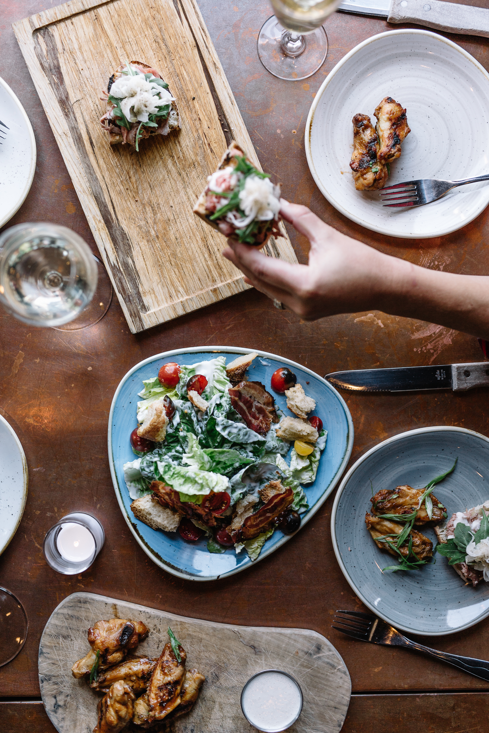 A table with various dishes including a salad, bread, grilled chicken wings, and small plates, along with glasses of white wine and a lit candle.