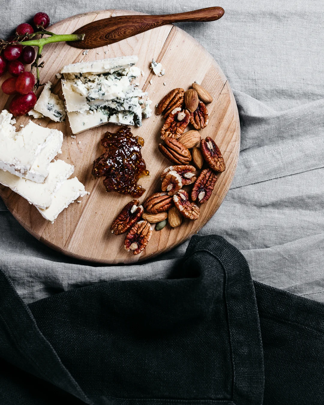 Cheese, nuts, grapes, and jam on a round wooden cheese board.