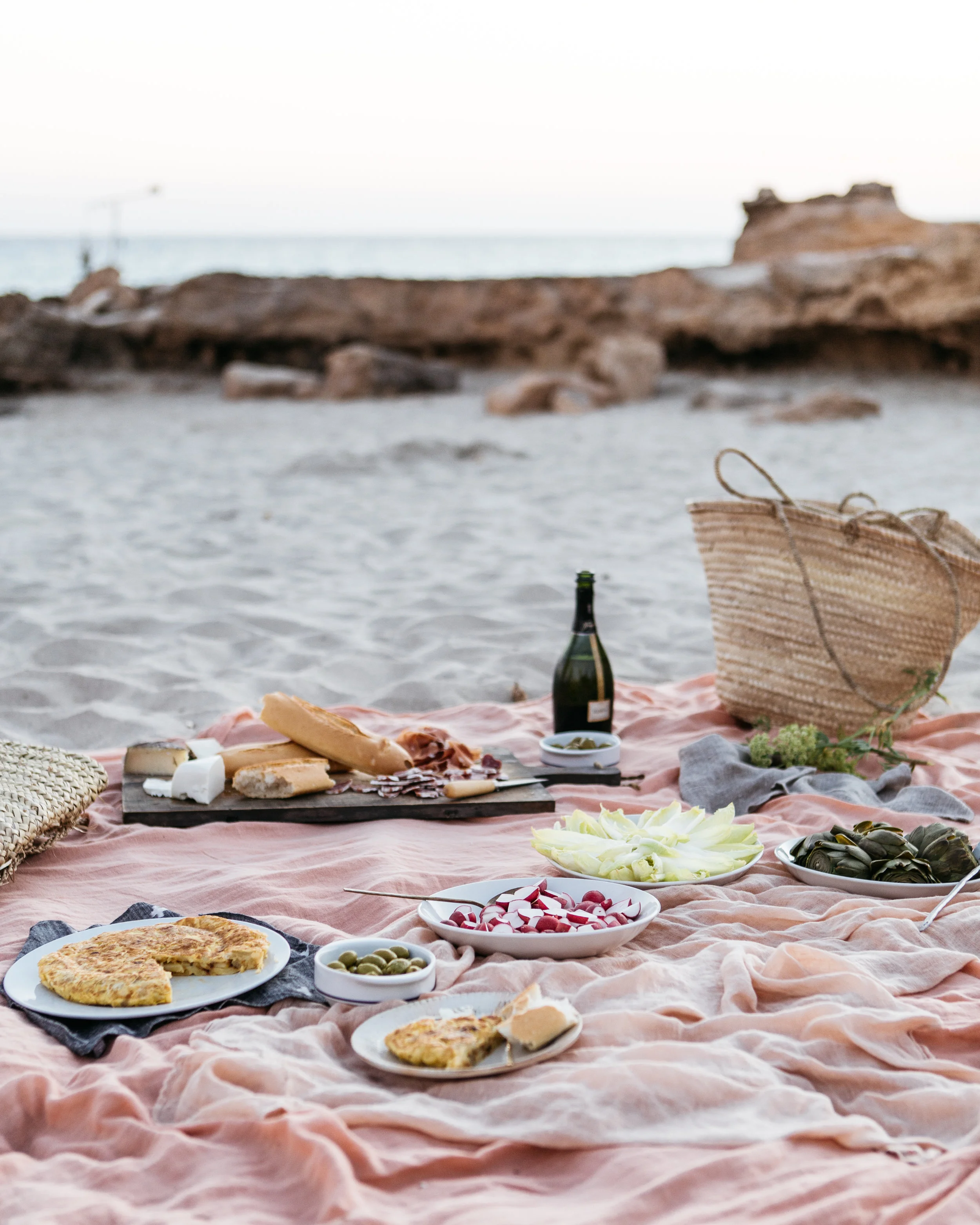 A beach picnic setup with various dishes including a quiche, radishes, green olives, and cheese, along with a bottle of wine, a basket, and the ocean in the background.