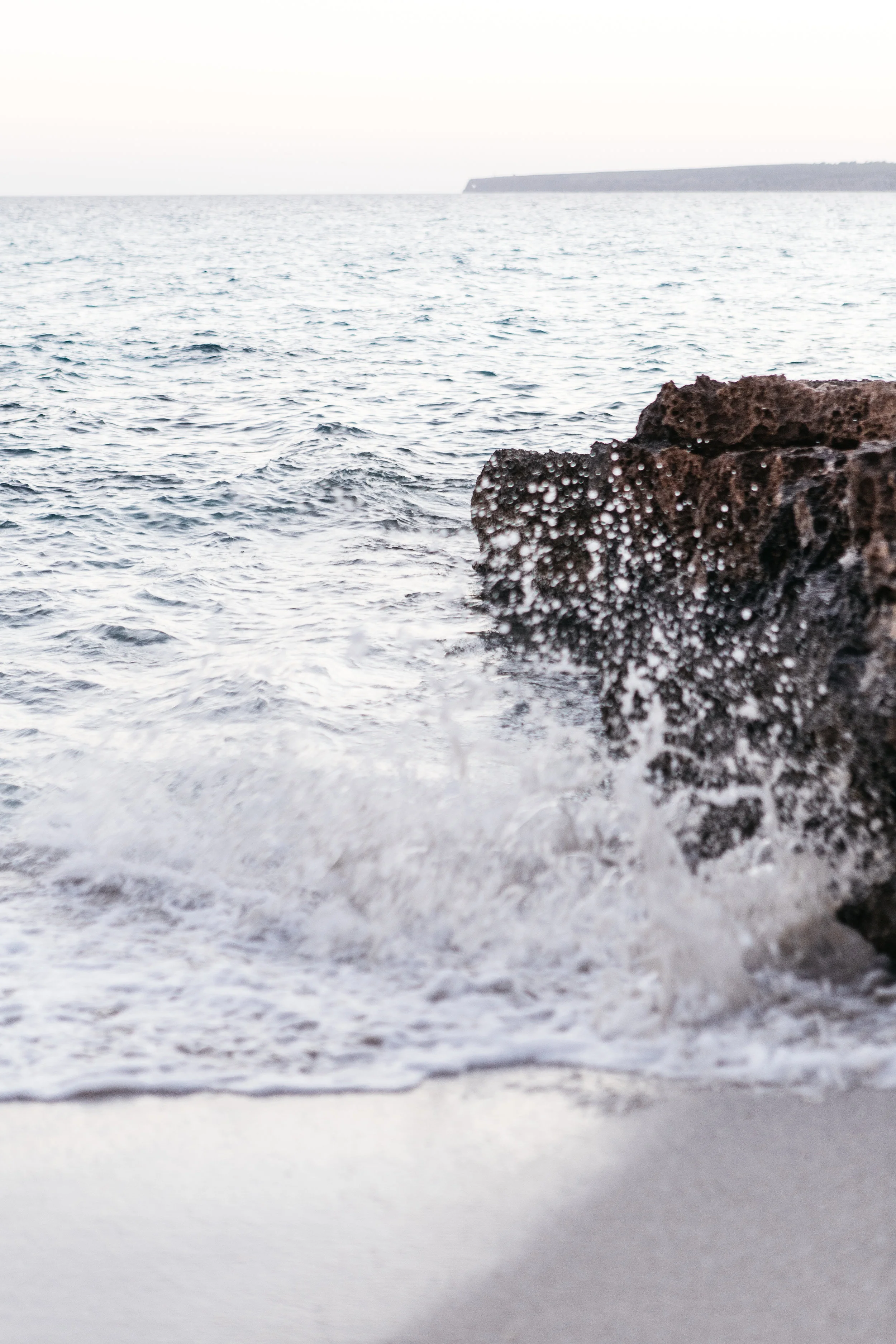 Ocean waves hitting rocks on a beach with distant cliffs in the background during sunset or sunrise.