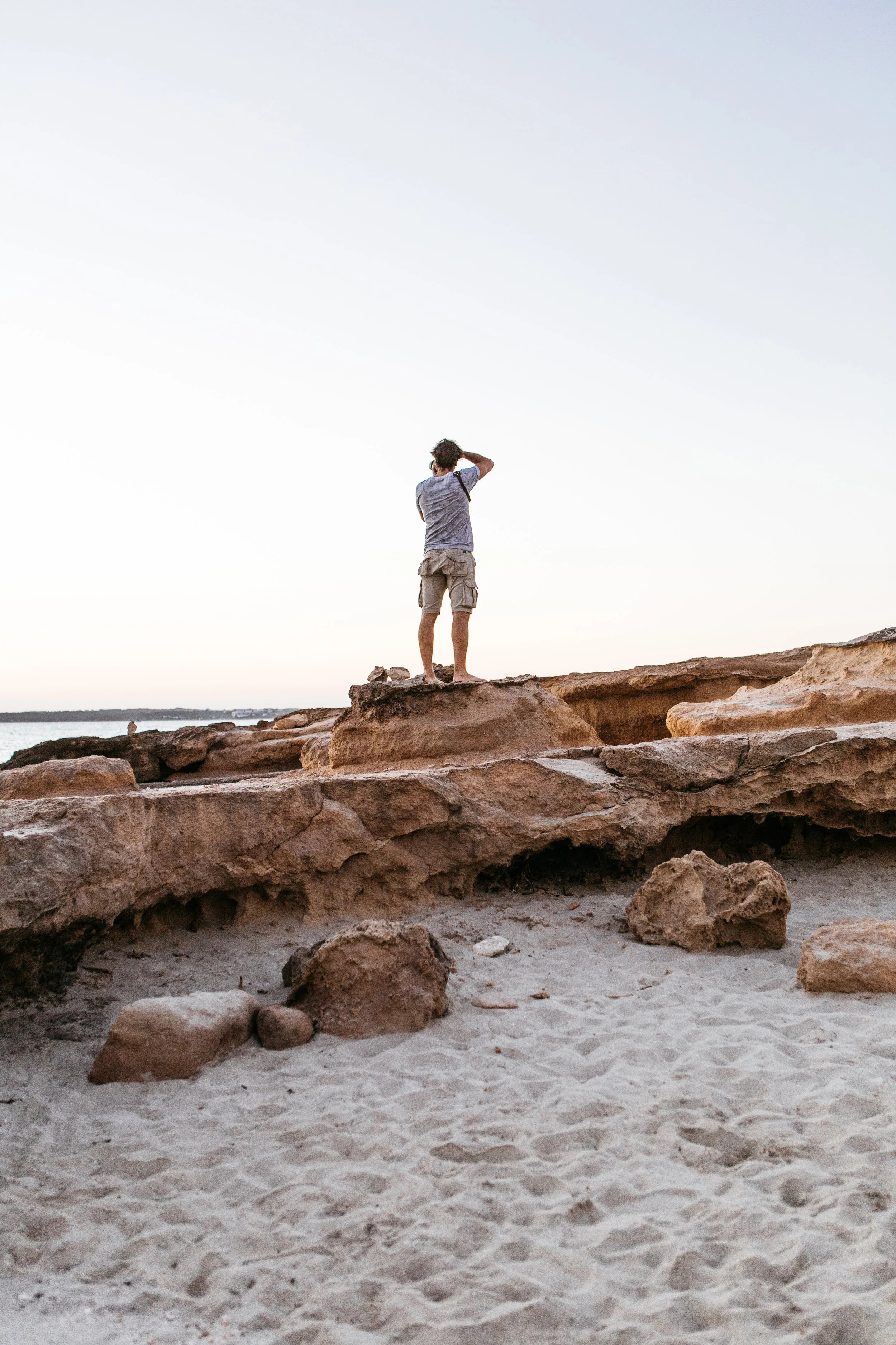 Person standing on rocks at the beach, looking out at the water during sunset or sunrise.