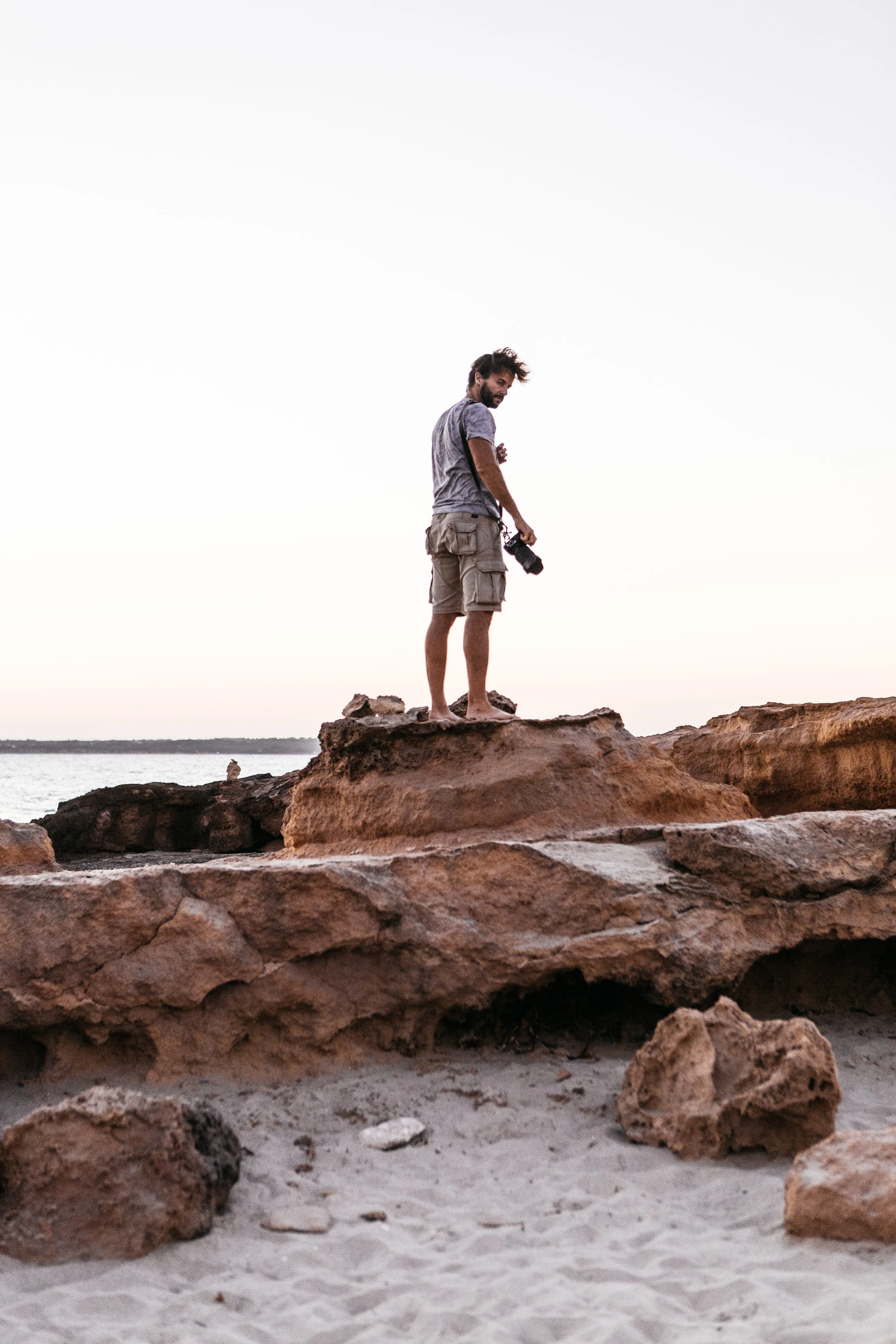 A man standing barefoot on rocky terrain by the water, holding a camera, during sunset or sunrise.