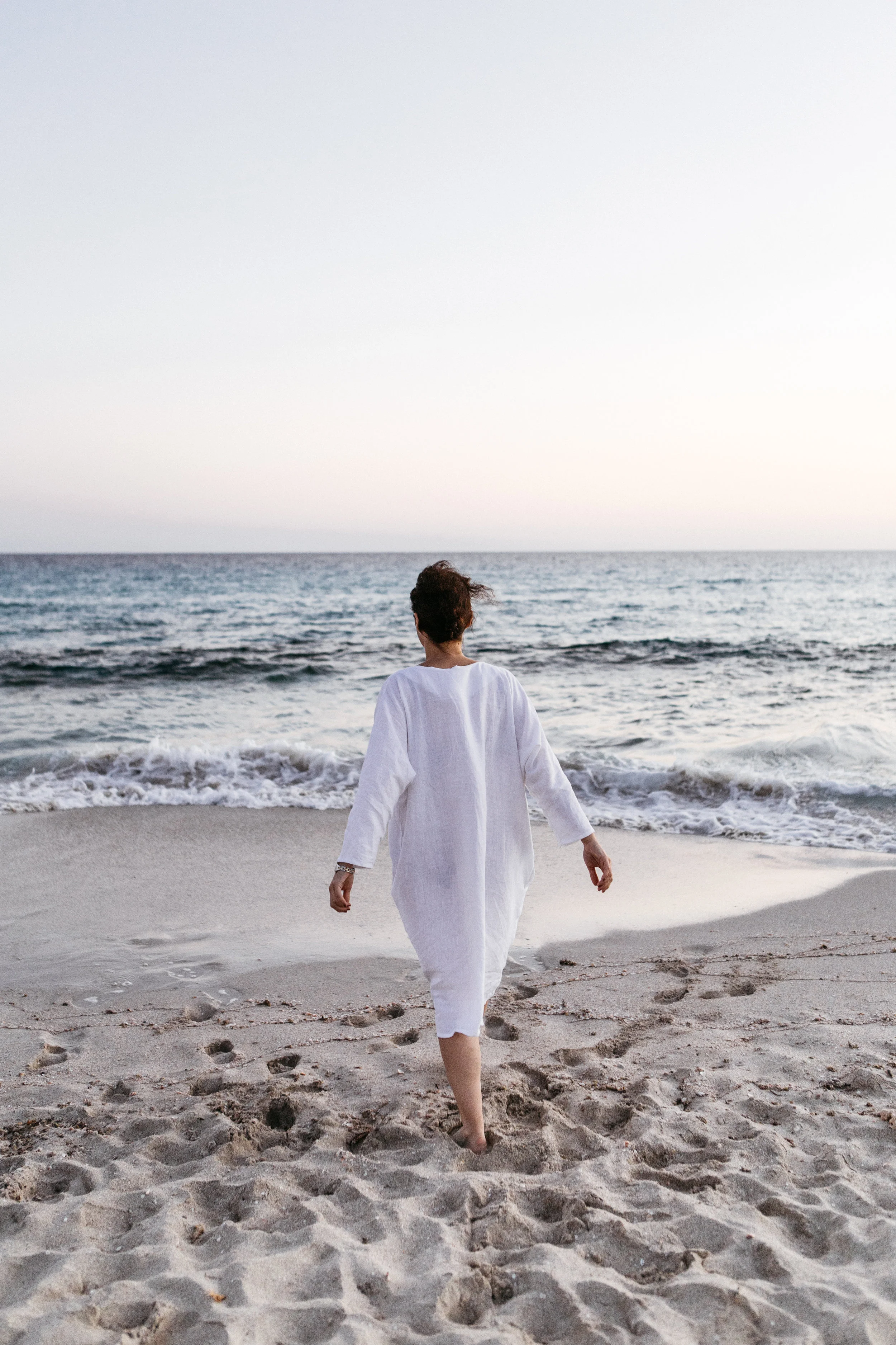 Person in a white dress walking towards the ocean on a sandy beach at sunset.