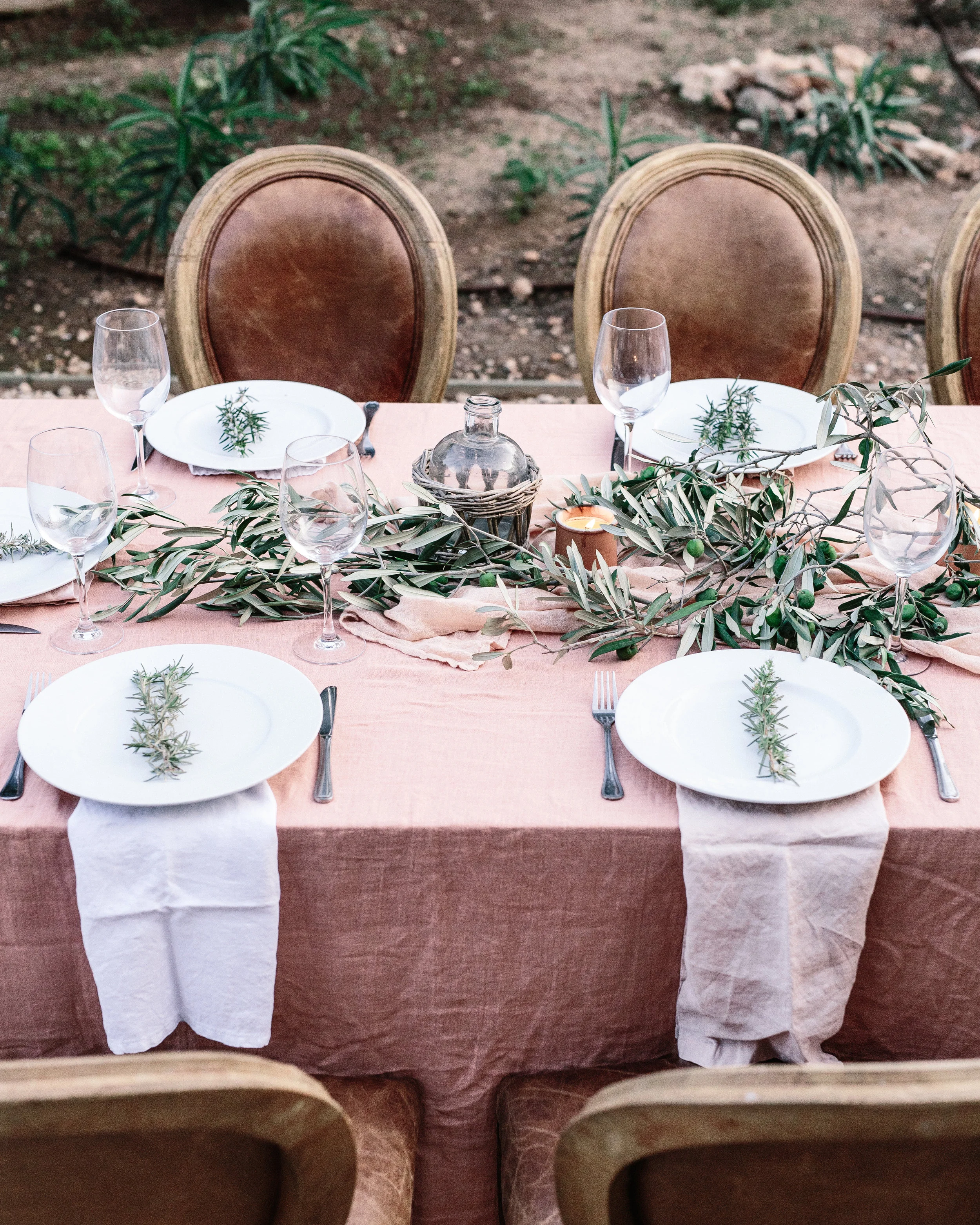 A beautifully set outdoor dining table with a pink linen tablecloth, white plates, silverware, and wine glasses. Greenery and olive branches as centerpieces, with a small candle and decorative bottle in the middle. Brown leather chairs surround the t