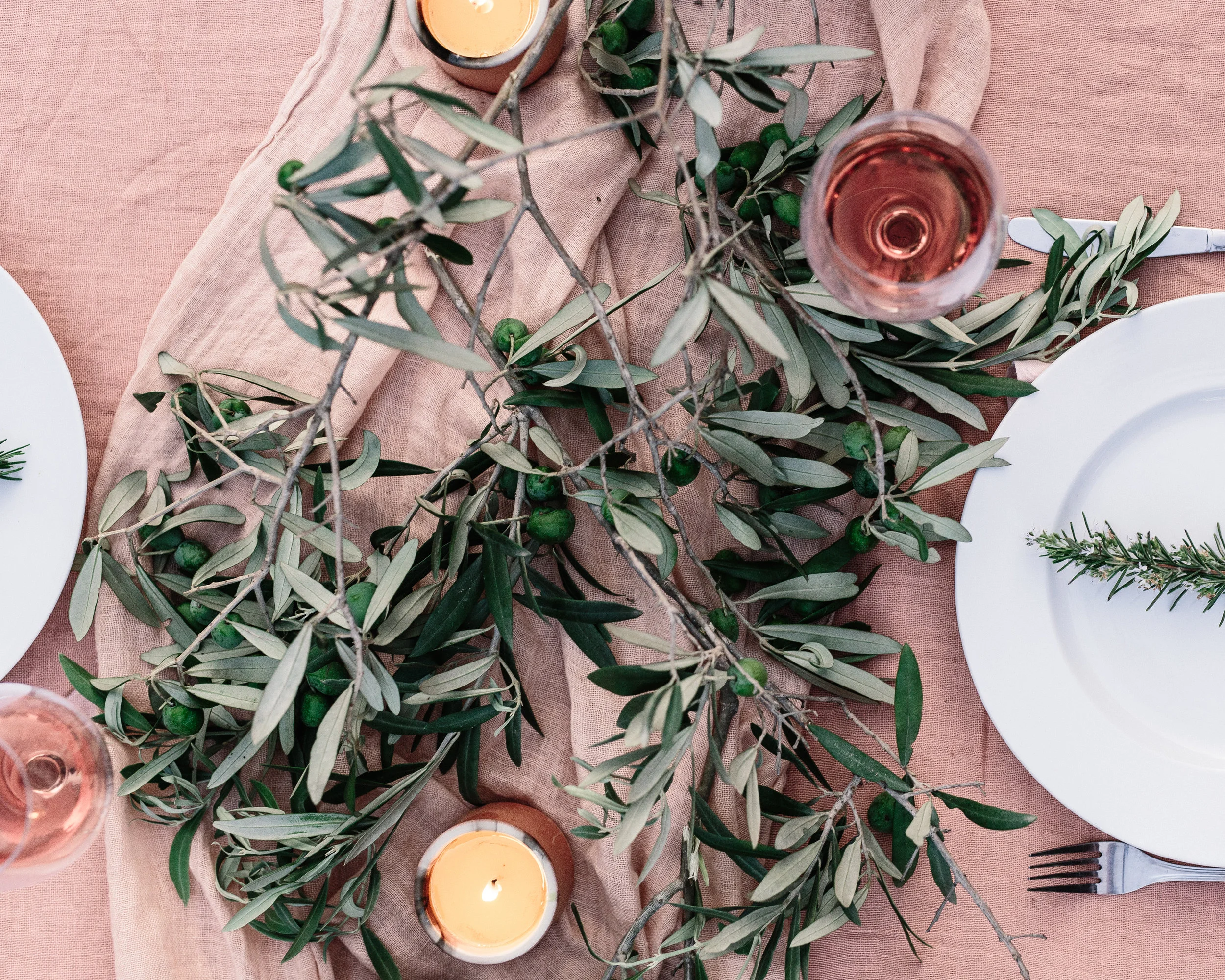 A table setting with a pink tablecloth, white plates, silverware, a branch of greenery and olives, and lit candles.