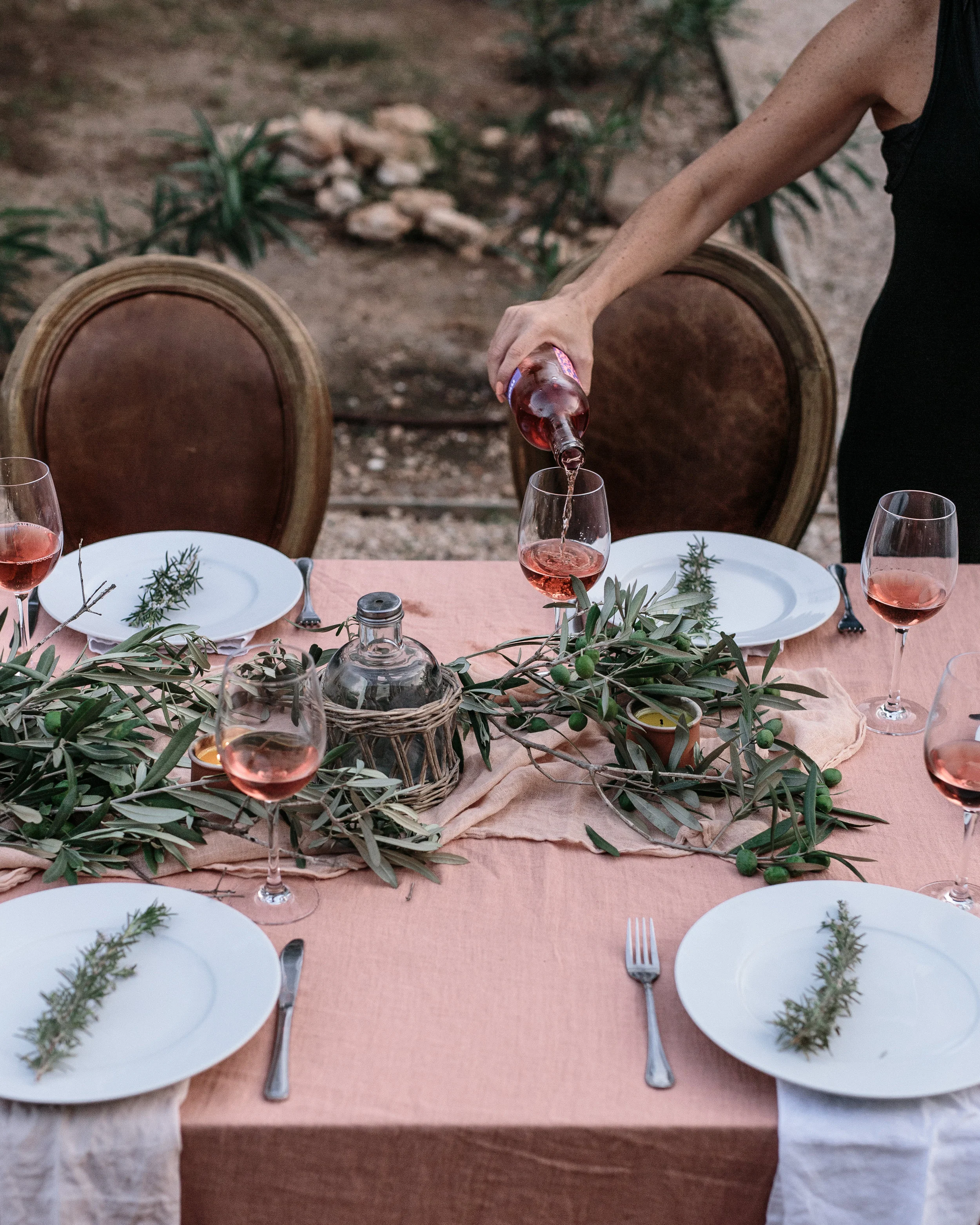 A person pouring rosé wine into a glass on a decorated outdoor table with plates, silverware, and greenery.