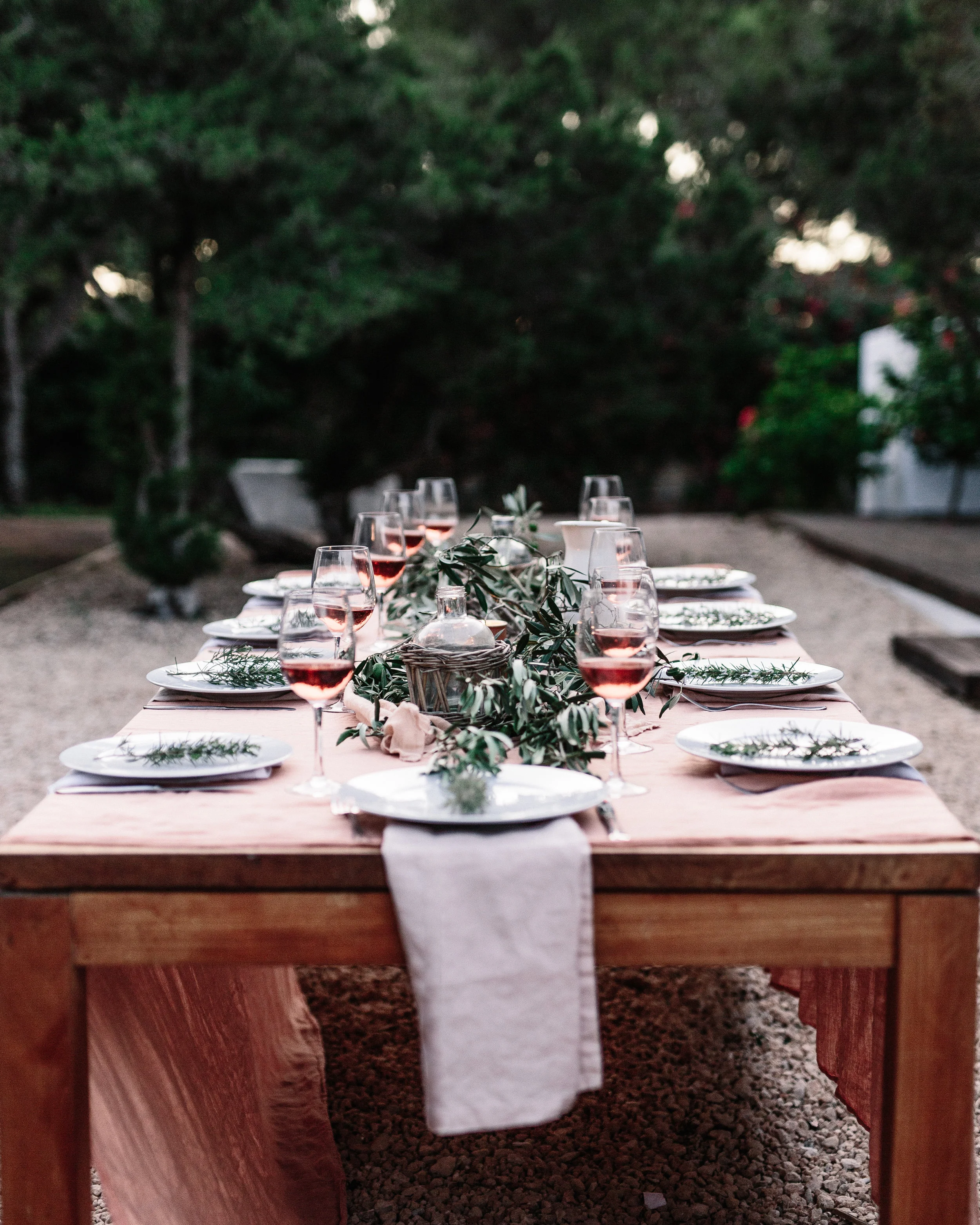 Outdoor dining table set for a meal with wine glasses, white plates, green foliage, and a linen table runner, surrounded by trees.