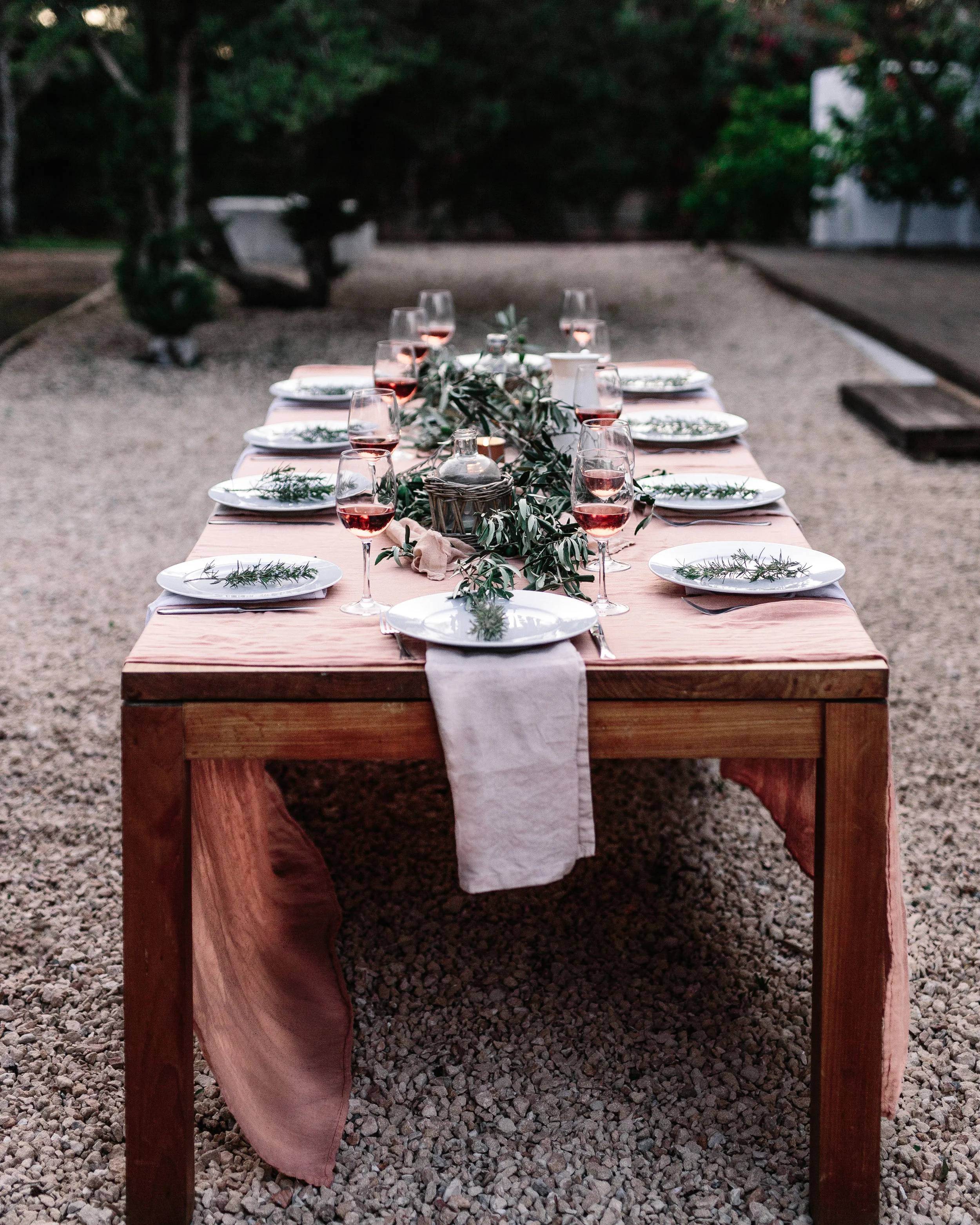 A long outdoor wooden dining table set for a meal, decorated with green foliage, white plates, wine glasses filled with pinkish red wine, and a beige table runner, situated on a gravel surface with a background of trees and outdoor scenery.