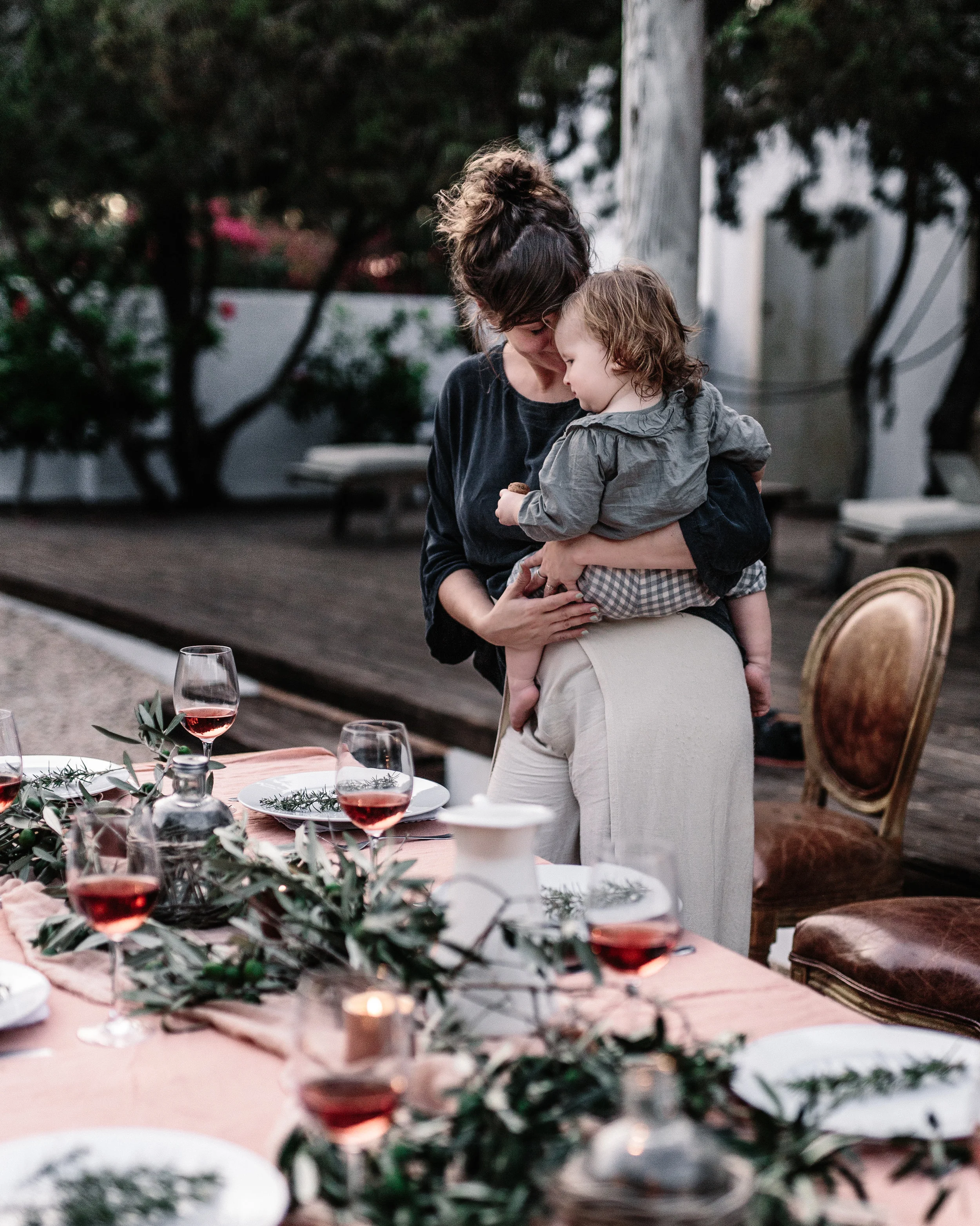 A woman holding a young child close during an outdoor dinner party with a decorated table of wine glasses, plates, and greenery.