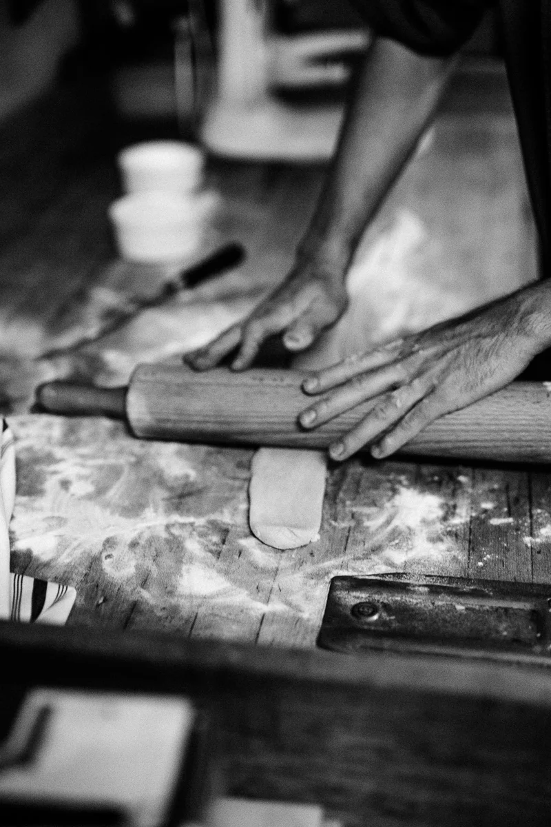 Hands of a person using a rolling pin to flatten dough on a floured wooden surface, with bowls and a spatula in the background.