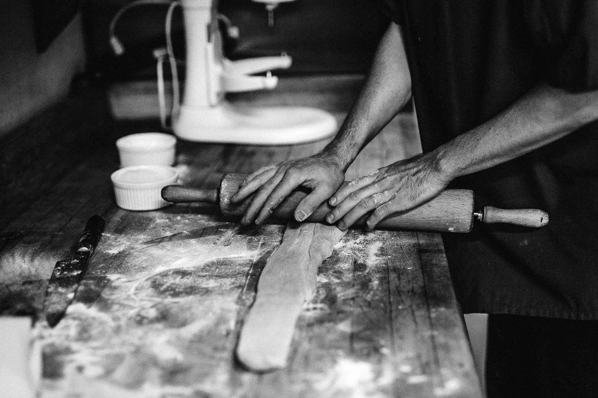 Person rolling out dough with a rolling pin on a kitchen counter, surrounded by baking tools and ingredients, in black and white.