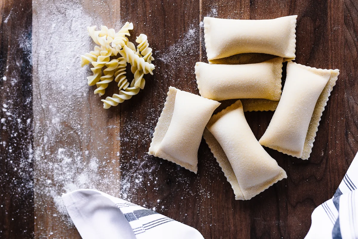 Uncooked ravioli and rotini pasta on a wooden surface dusted with flour, with a striped cloth napkin in the corner.