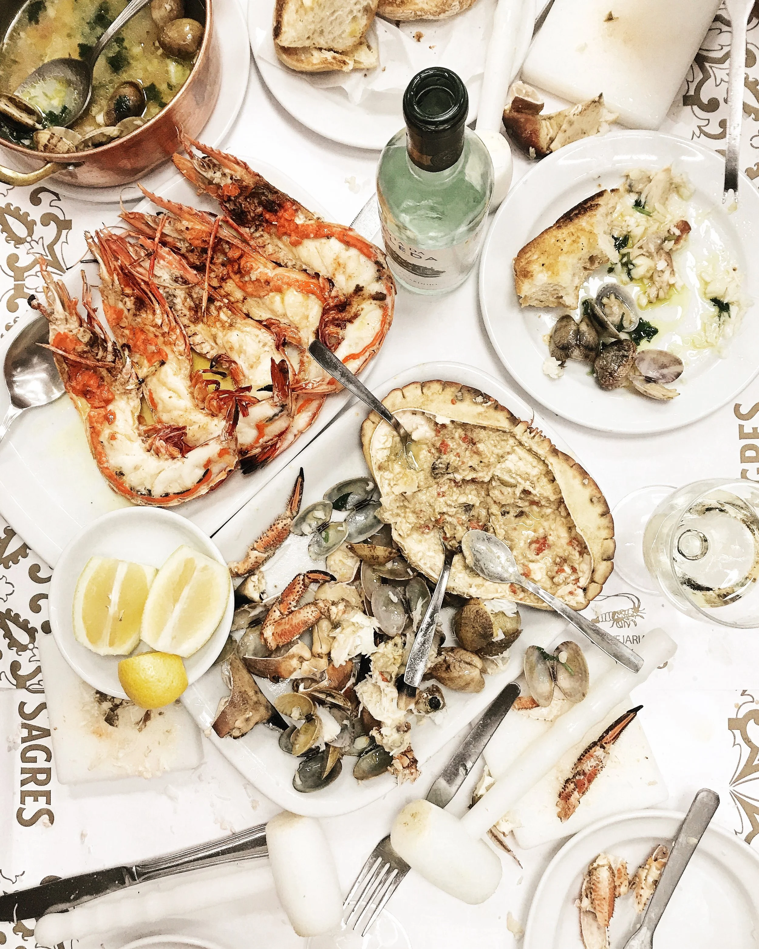 Seafood feast with grilled prawns, clams, crab, and lobster, with lemon wedges, bread, and wine on the table.