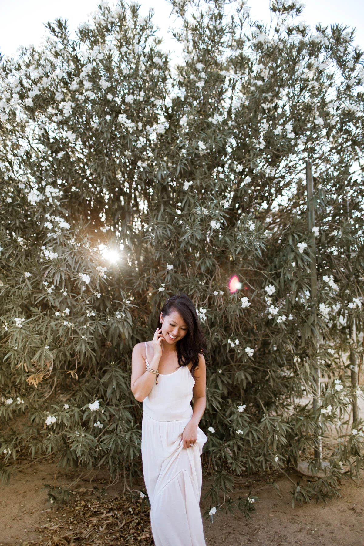 A woman in a white dress standing outdoors near flowering shrub, smiling and looking down, with sunlight shining through the shrub.