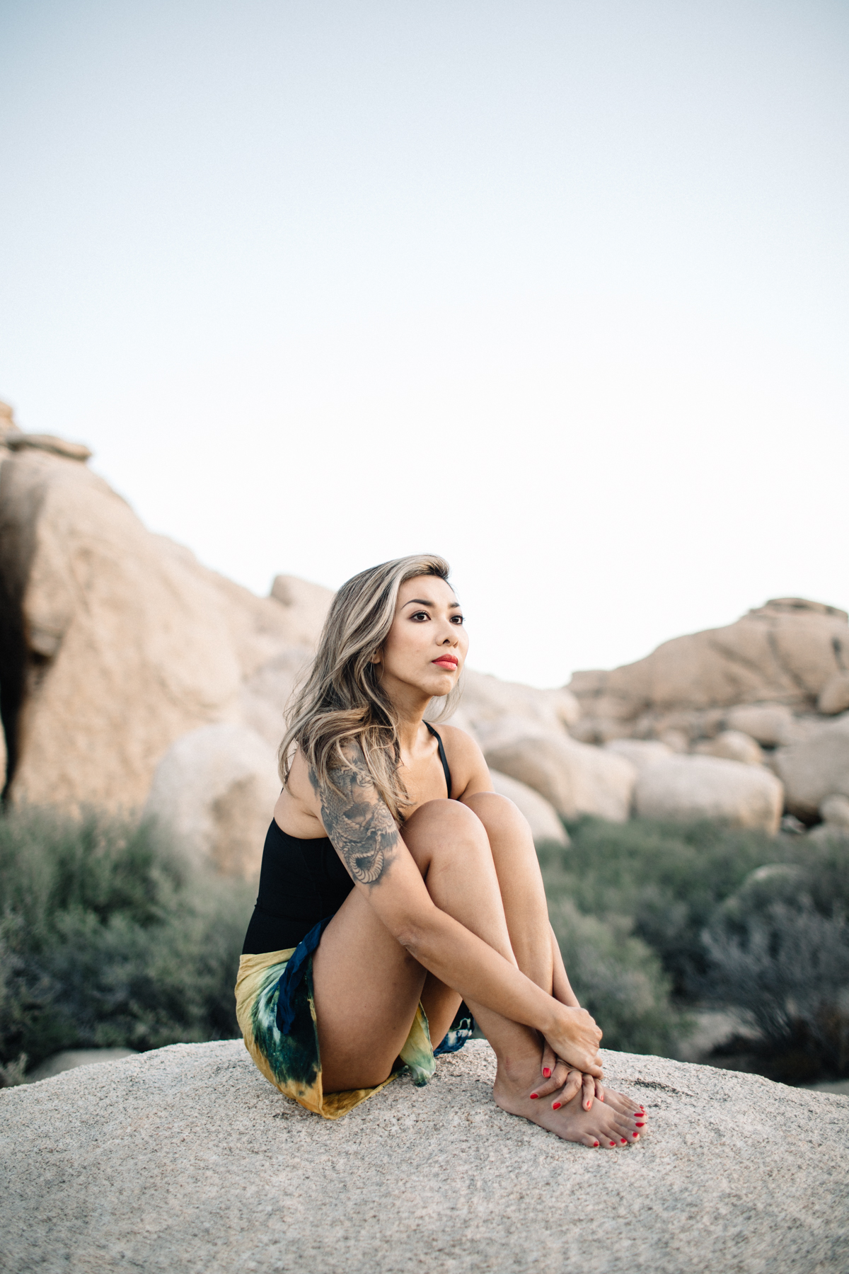 A woman with blonde hair and a tattoo on her arm sitting on a rock in a desert landscape with large boulders and sparse vegetation, wearing a black tank top and patterned shorts.