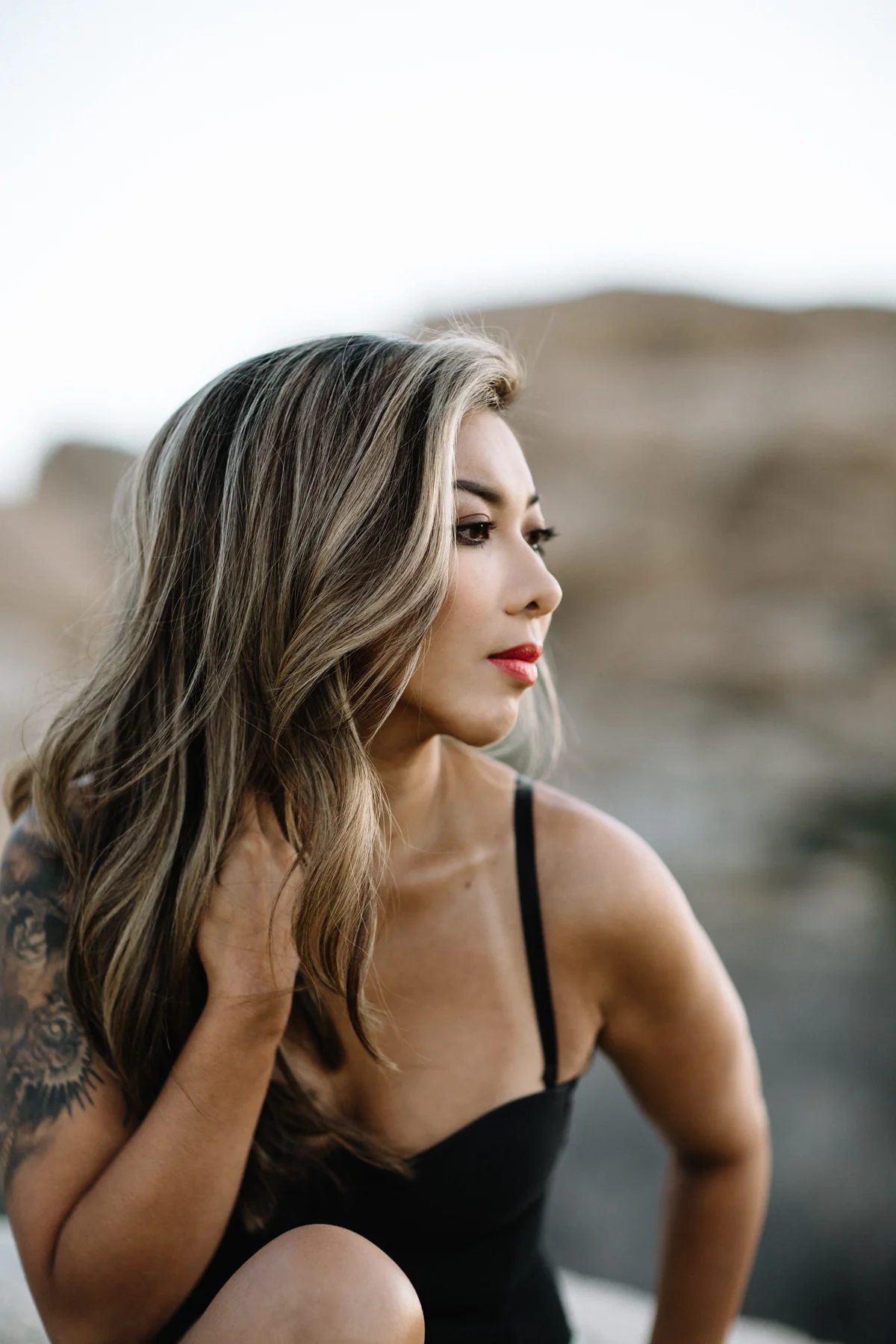 A woman with long, wavy hair and a tattoo on her arm, wearing a black tank top, looking to the side with a serious expression outdoors.