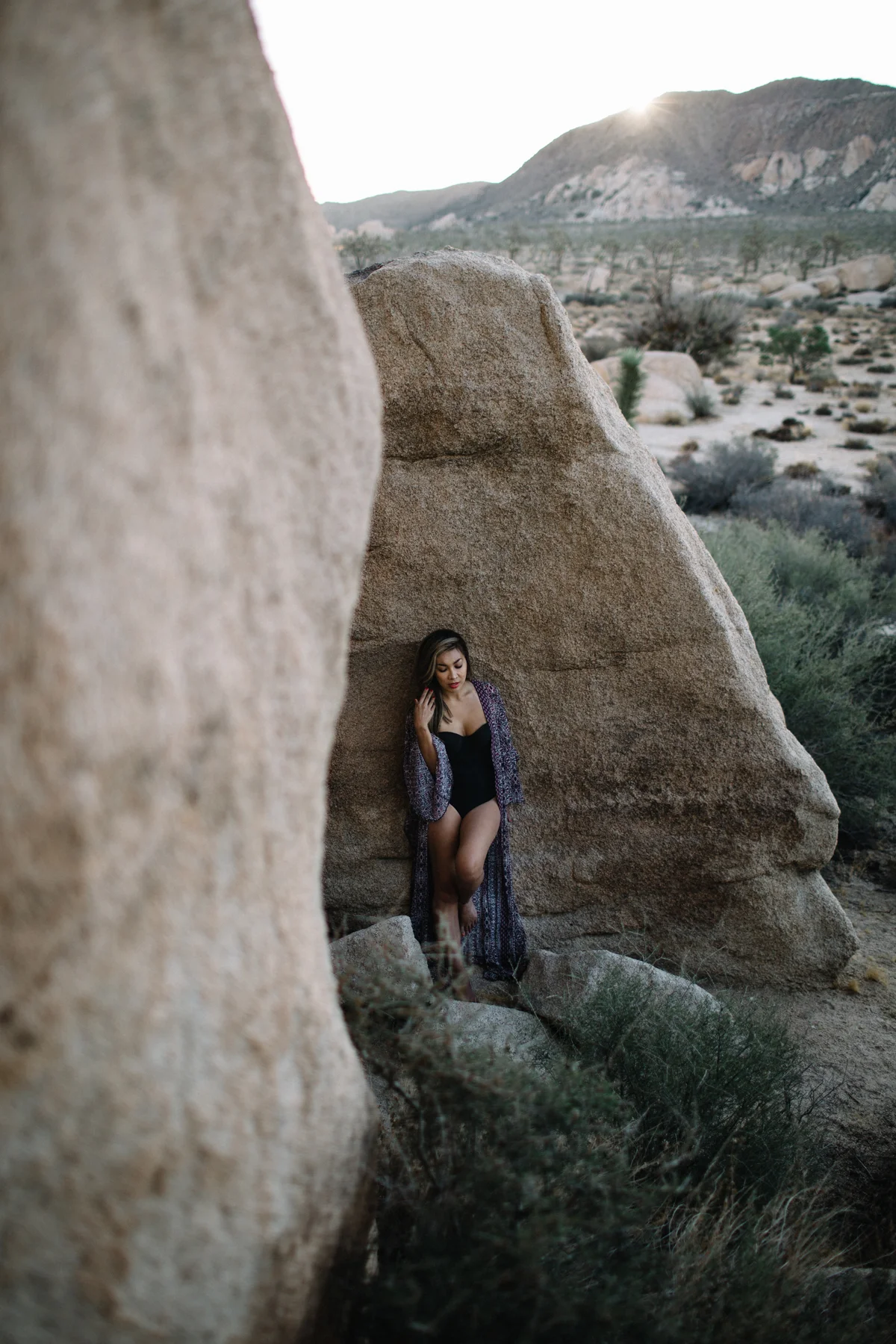 A woman in a black swimsuit and robe standing between large desert rocks, with a desert landscape and mountains in the background, at sunset.