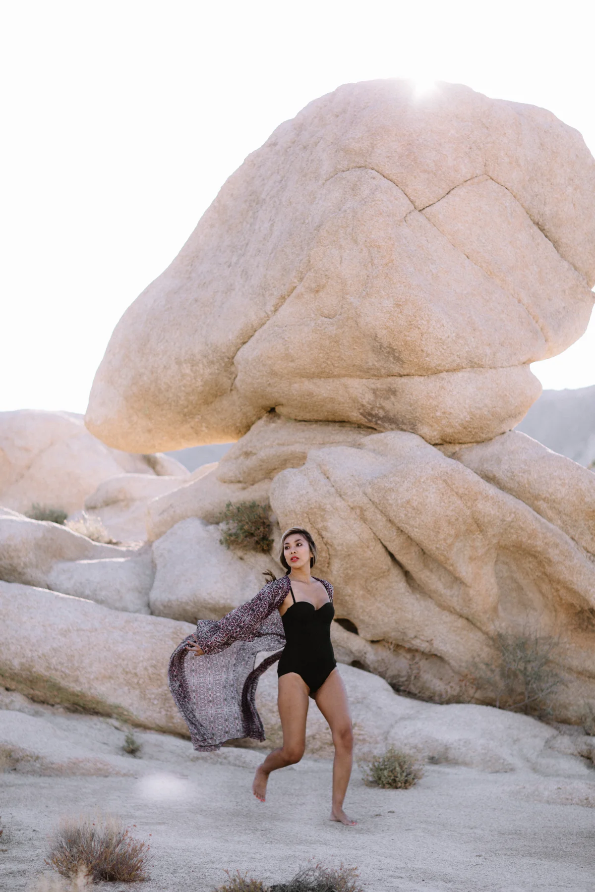A woman in a black swimsuit and a patterned cover-up walking barefoot in a desert landscape with large rock formations.