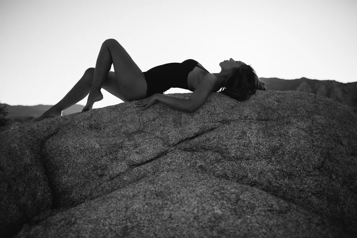 A woman lying on a large rock outdoors, wearing a black swimsuit, with her eyes closed and head tilted back.