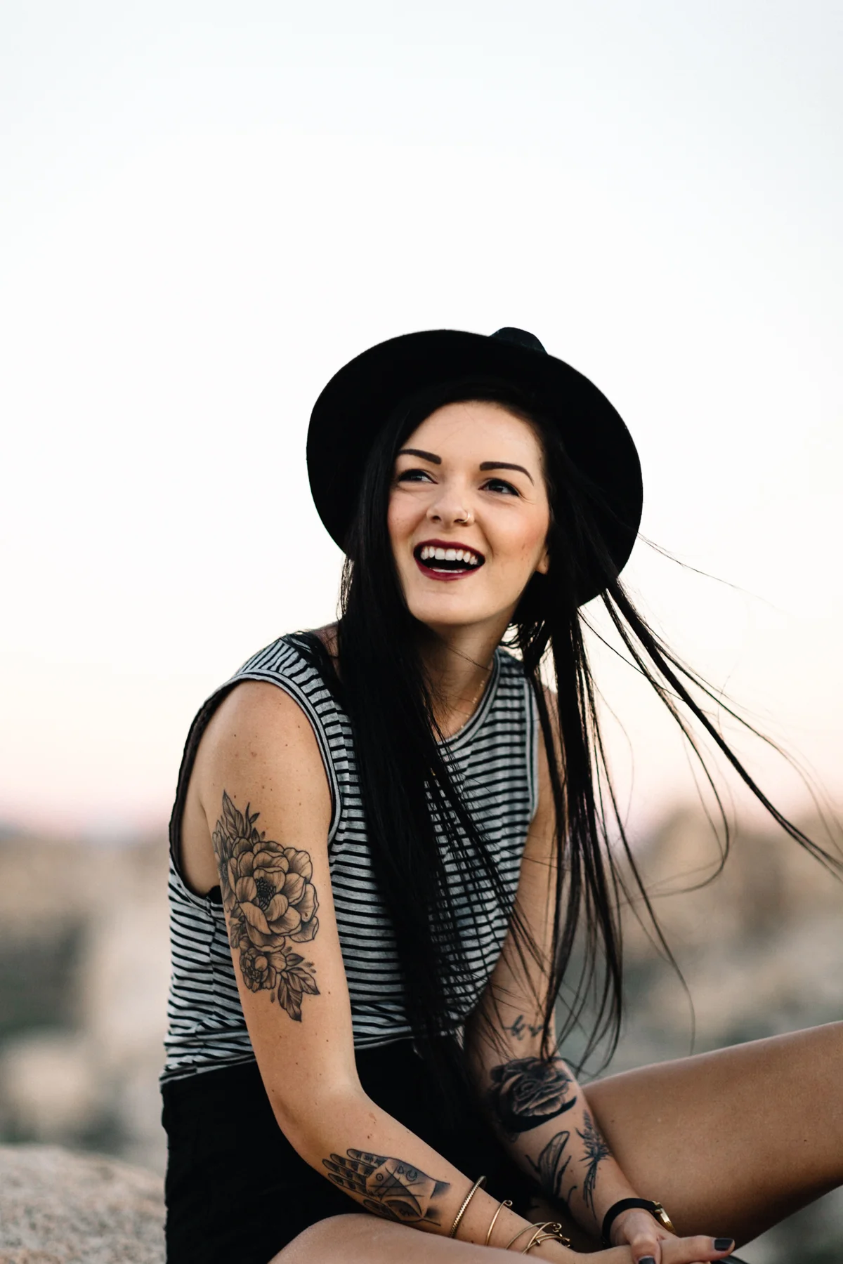 A young woman with long black hair, wearing a black hat and striped tank top, smiling outdoors during sunset with tattoos on her arm and wrist.