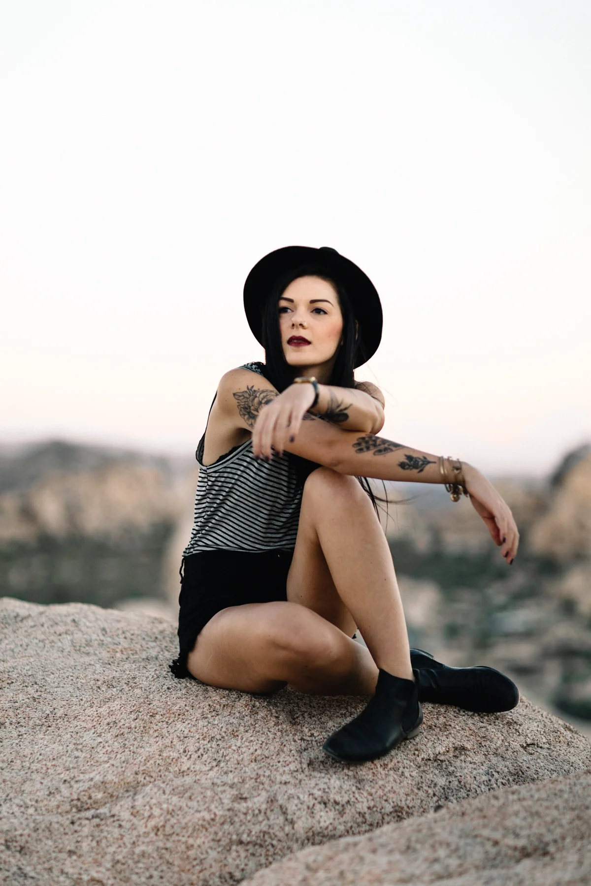 A woman with tattoos and dark hair sitting on a rock outdoors during sunset, wearing a black hat, striped shirt, black shorts, and black boots.