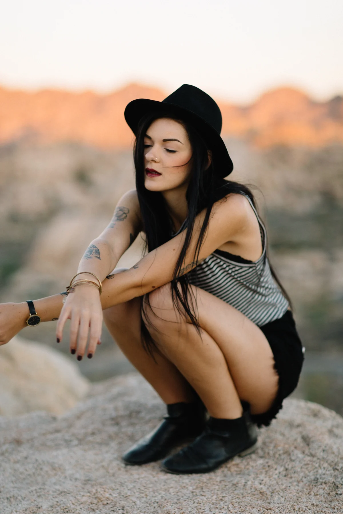A woman with long black hair, tattoos, and dark lipstick squats on a rock at sunset, wearing a black hat, striped tank top, and black shorts.