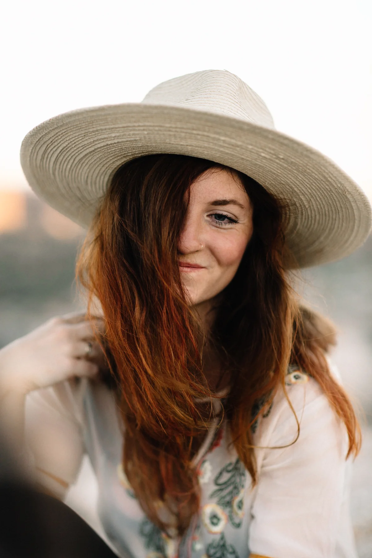A woman with long red hair wearing a wide-brimmed straw hat and a embroidered light-colored top outdoors.