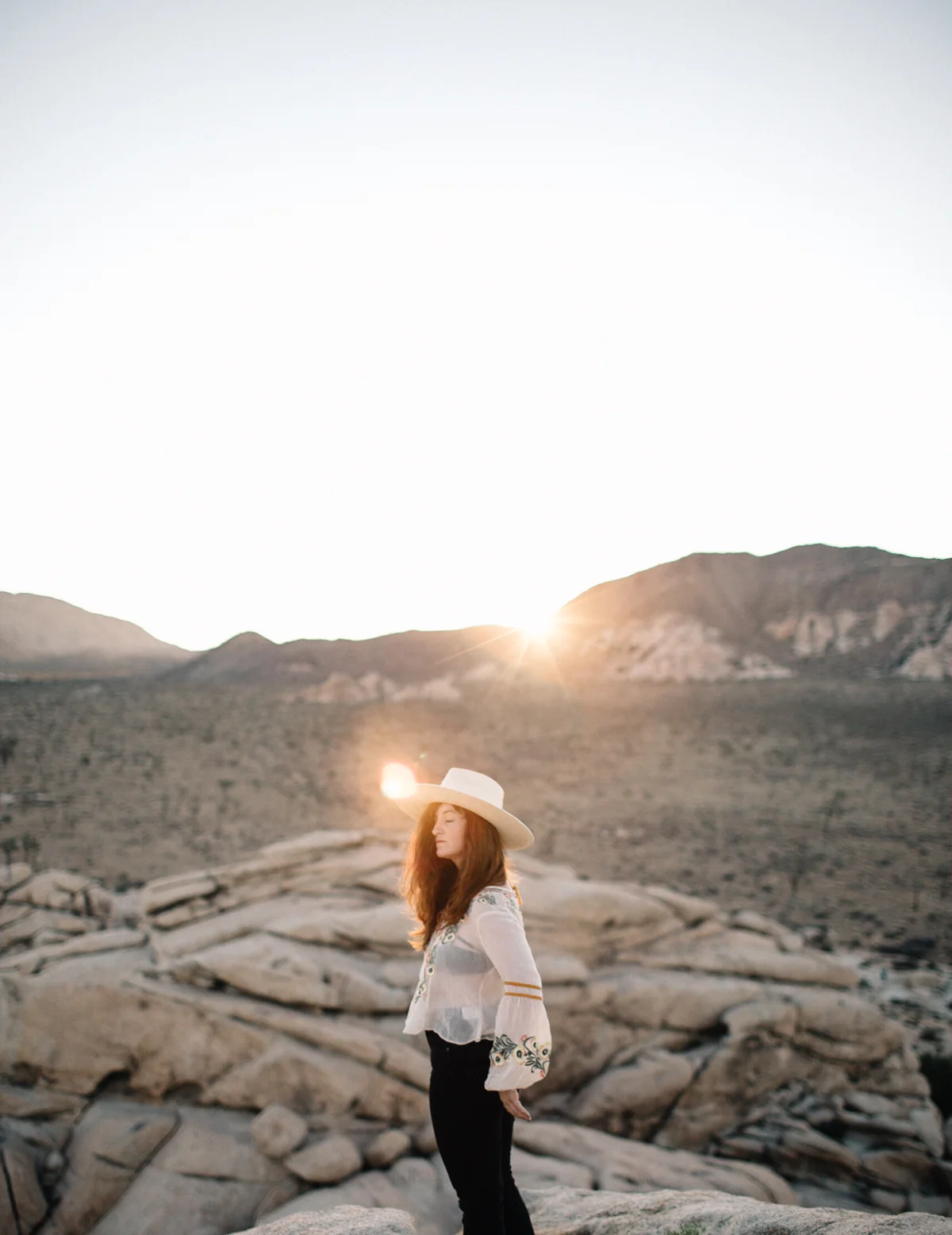 Woman standing on rocky terrain in desert at sunset, wearing a wide-brimmed hat and embroidered blouse.