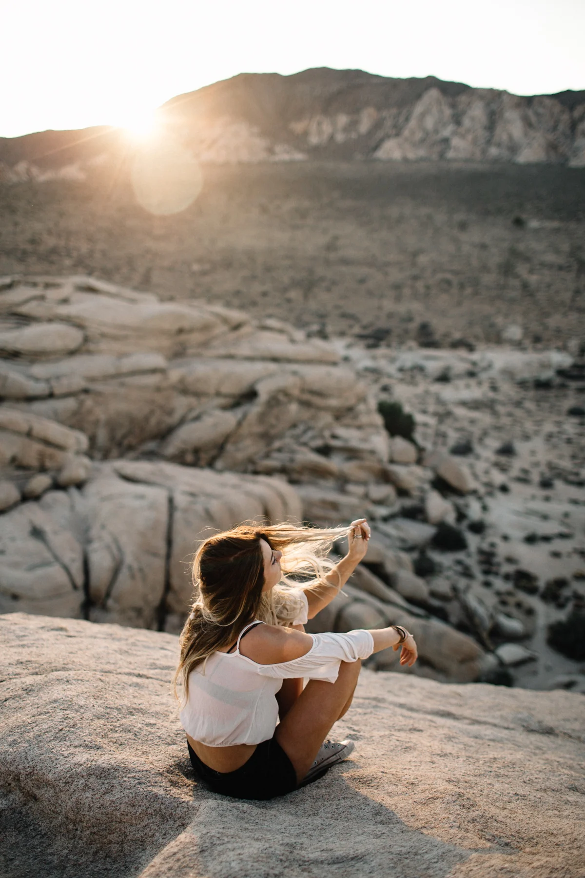 A woman sitting on a rock in a desert landscape, taking a selfie with the setting sun in the background.