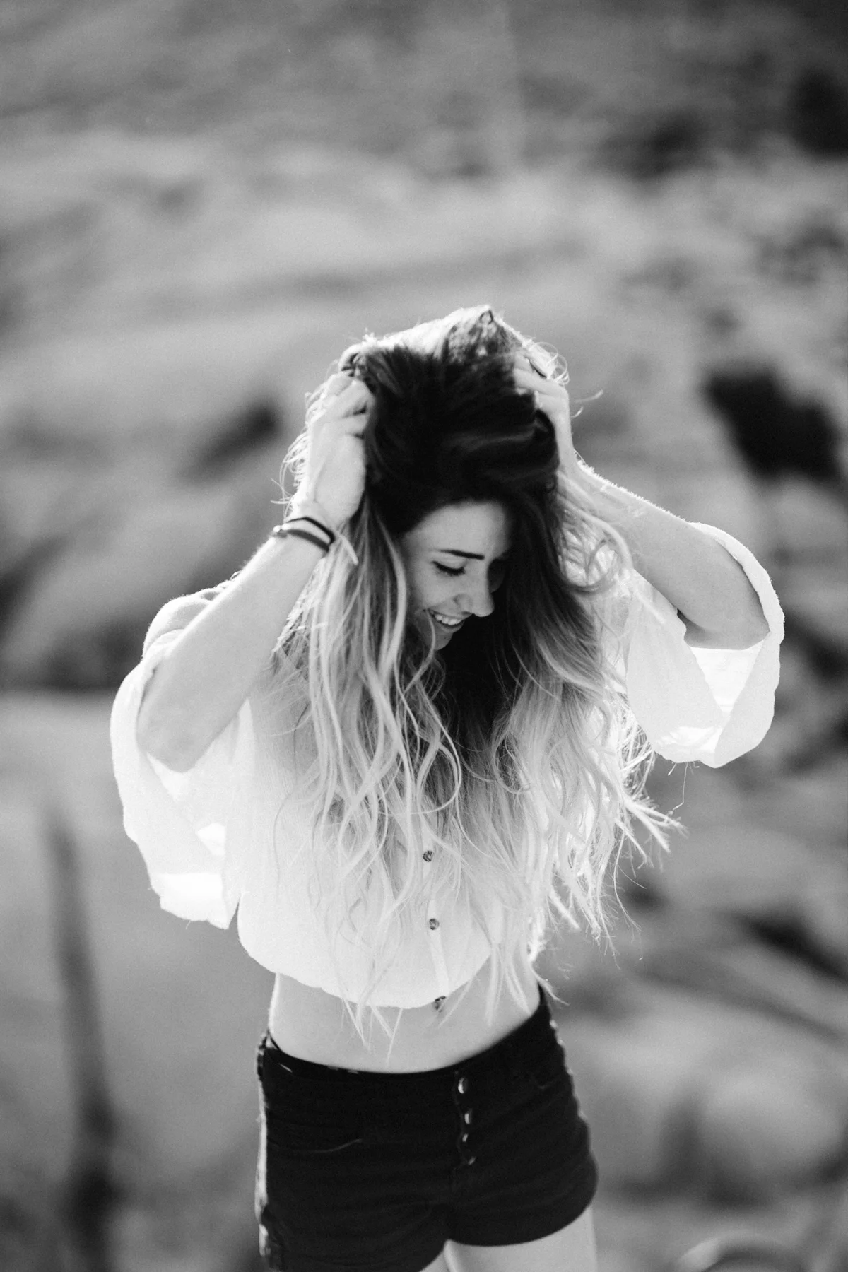 A woman with long, wavy hair, wearing a white blouse and black shorts, smiling and holding her head with both hands outdoors.