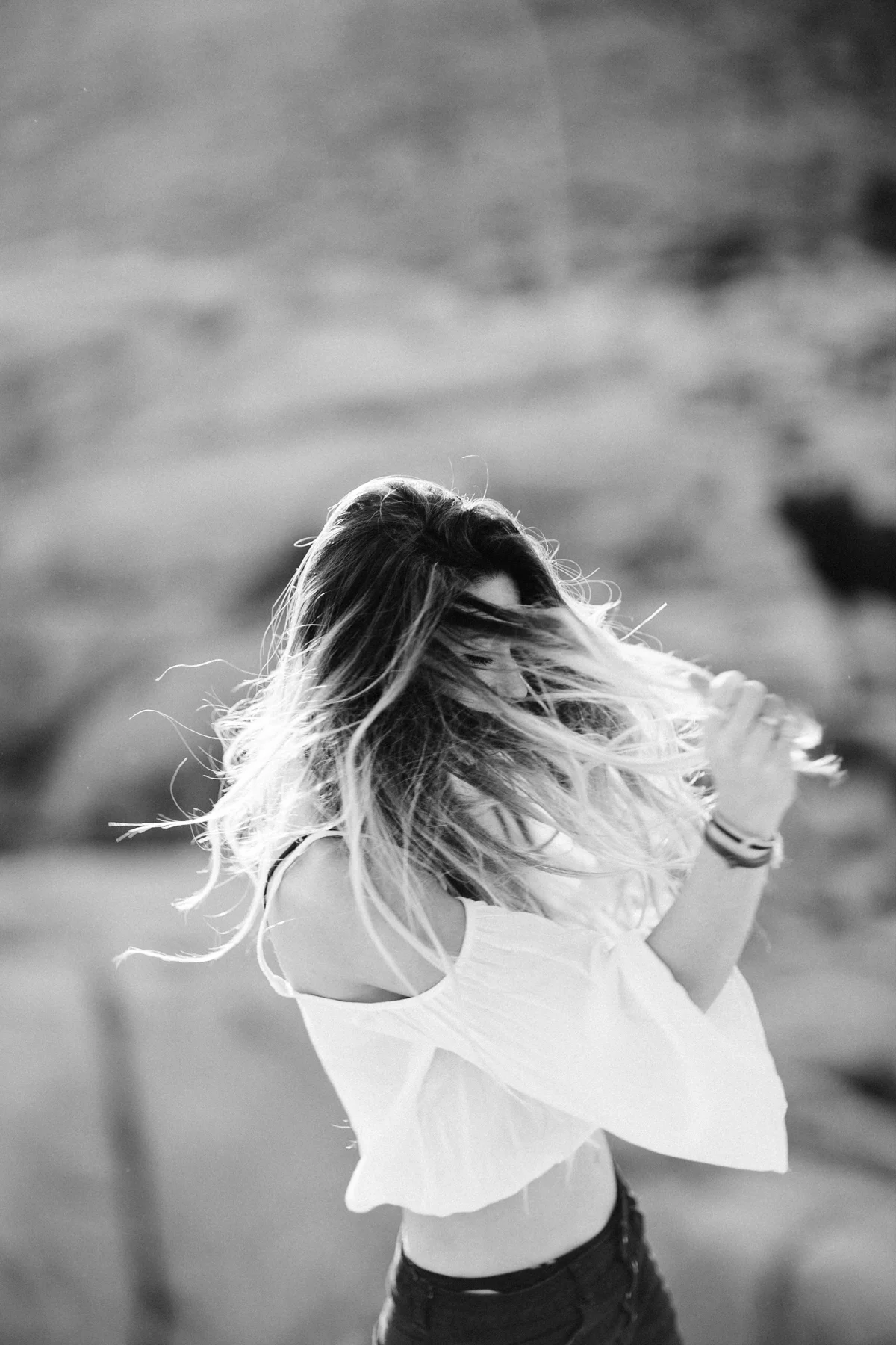 A woman with long hair, wearing a loose white top and dark pants, standing outdoors with her hair blowing in the wind.