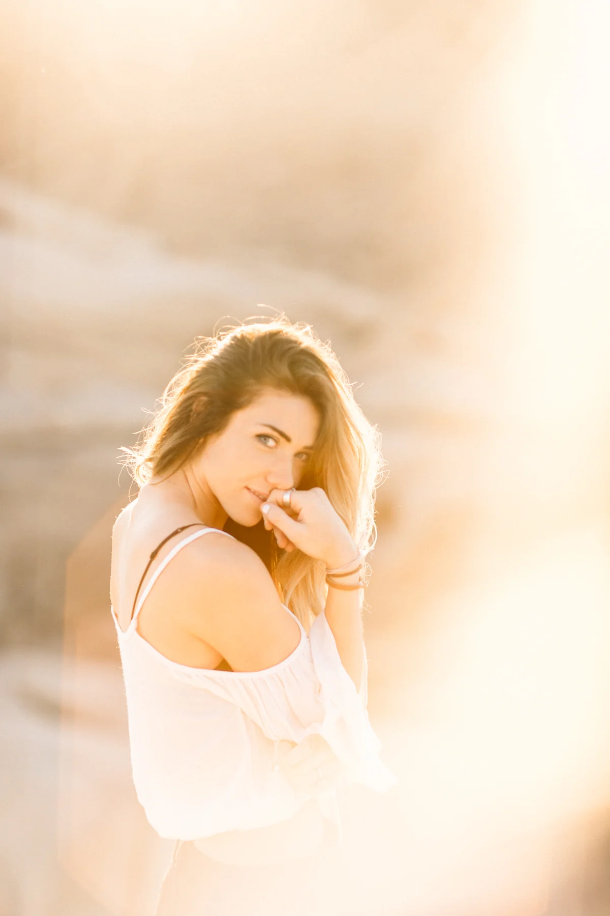 A young woman with long, wavy reddish hair stands outdoors at sunset, looking over her shoulder with a playful expression, wearing a white off-the-shoulder top.