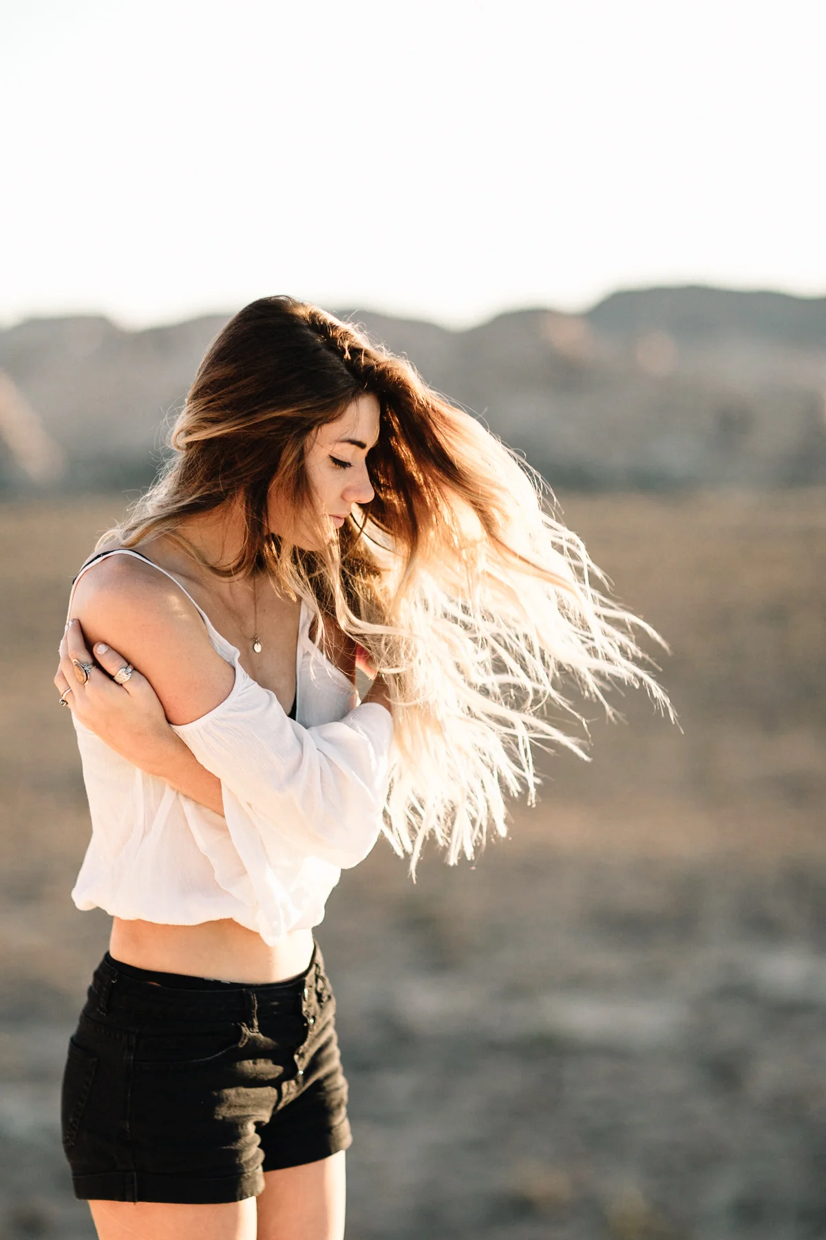A young woman with long, flowing hair stands outdoors in a natural setting during sunset, wearing a white off-shoulder top and black shorts, with her arms crossed over her chest and her eyes closed.
