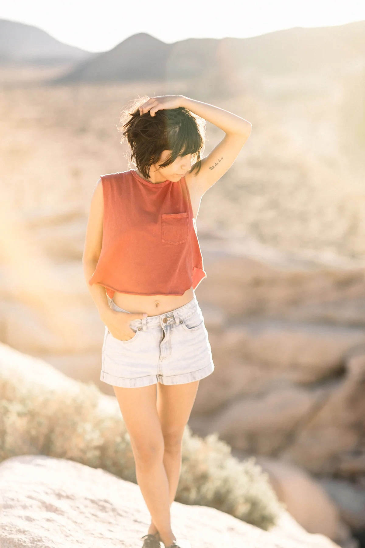 A young woman with short dark hair stands outdoors in a desert landscape during sunset, wearing a sleeveless red top, light denim shorts, and black shoes, looking down with her right hand touching her hair.