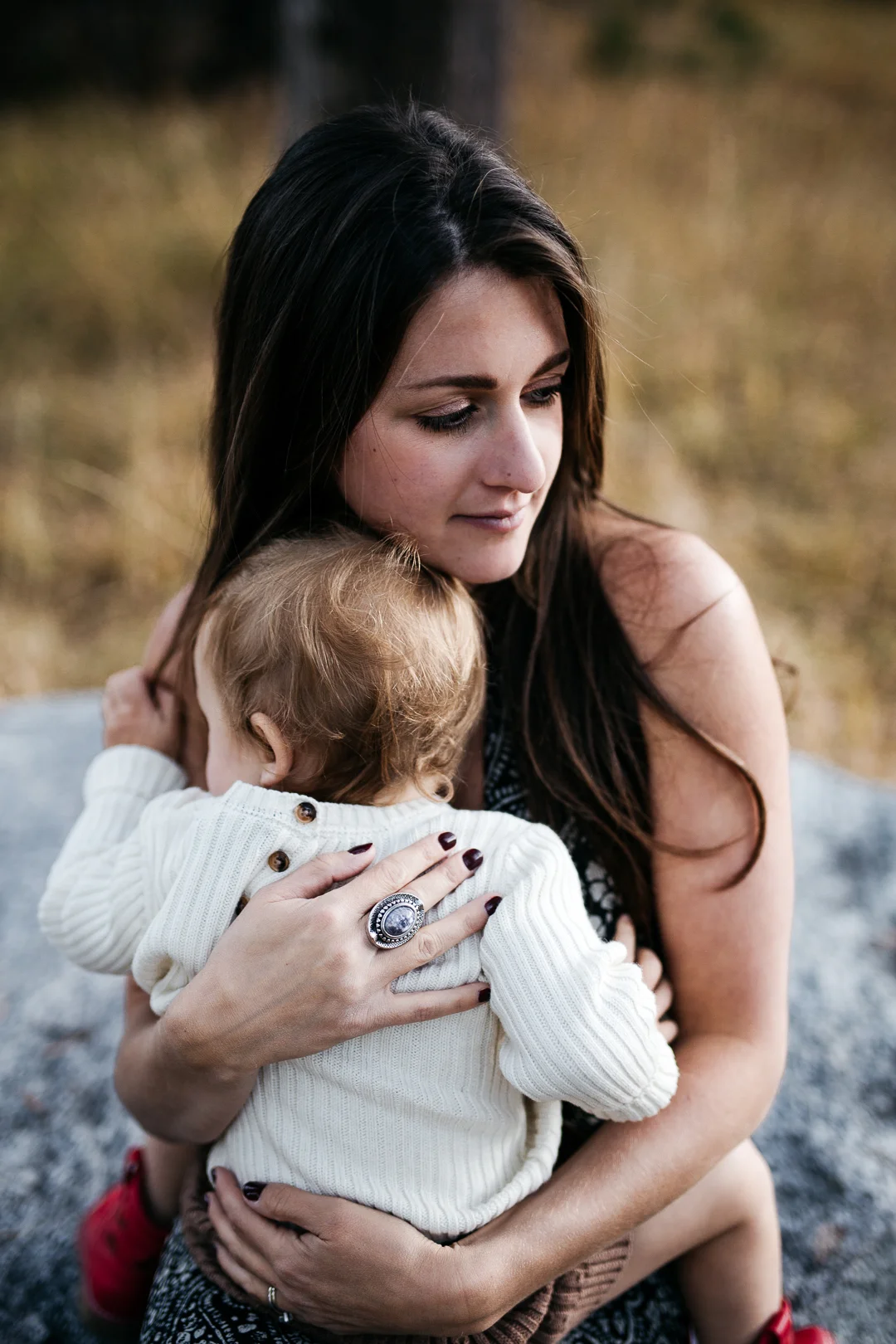 A woman with dark hair holding a small child with red hair, hugging in an outdoor setting with trees and grass in the background.