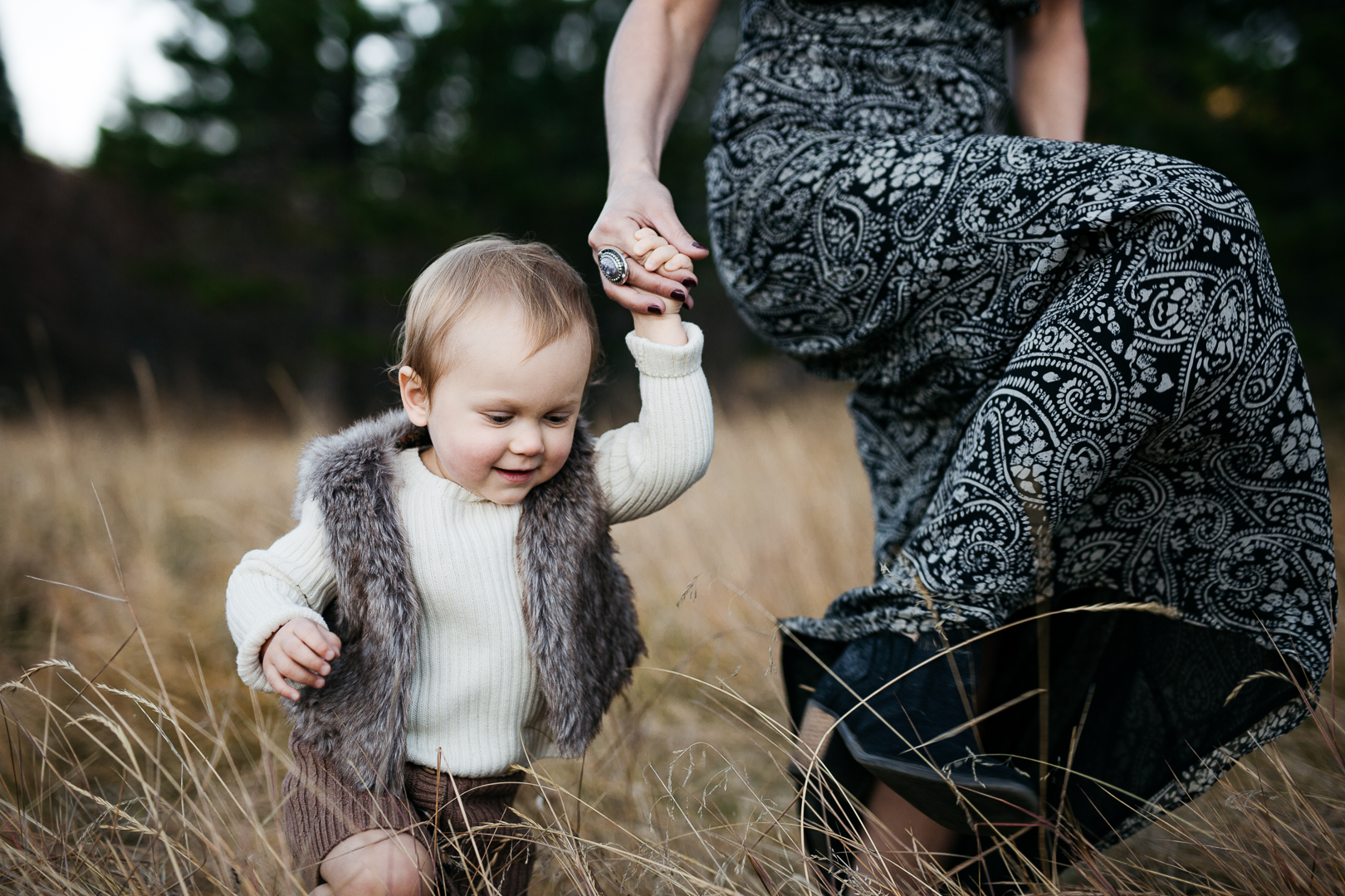 A woman holding a young child's hand as they walk through a field of tall grass outdoors.