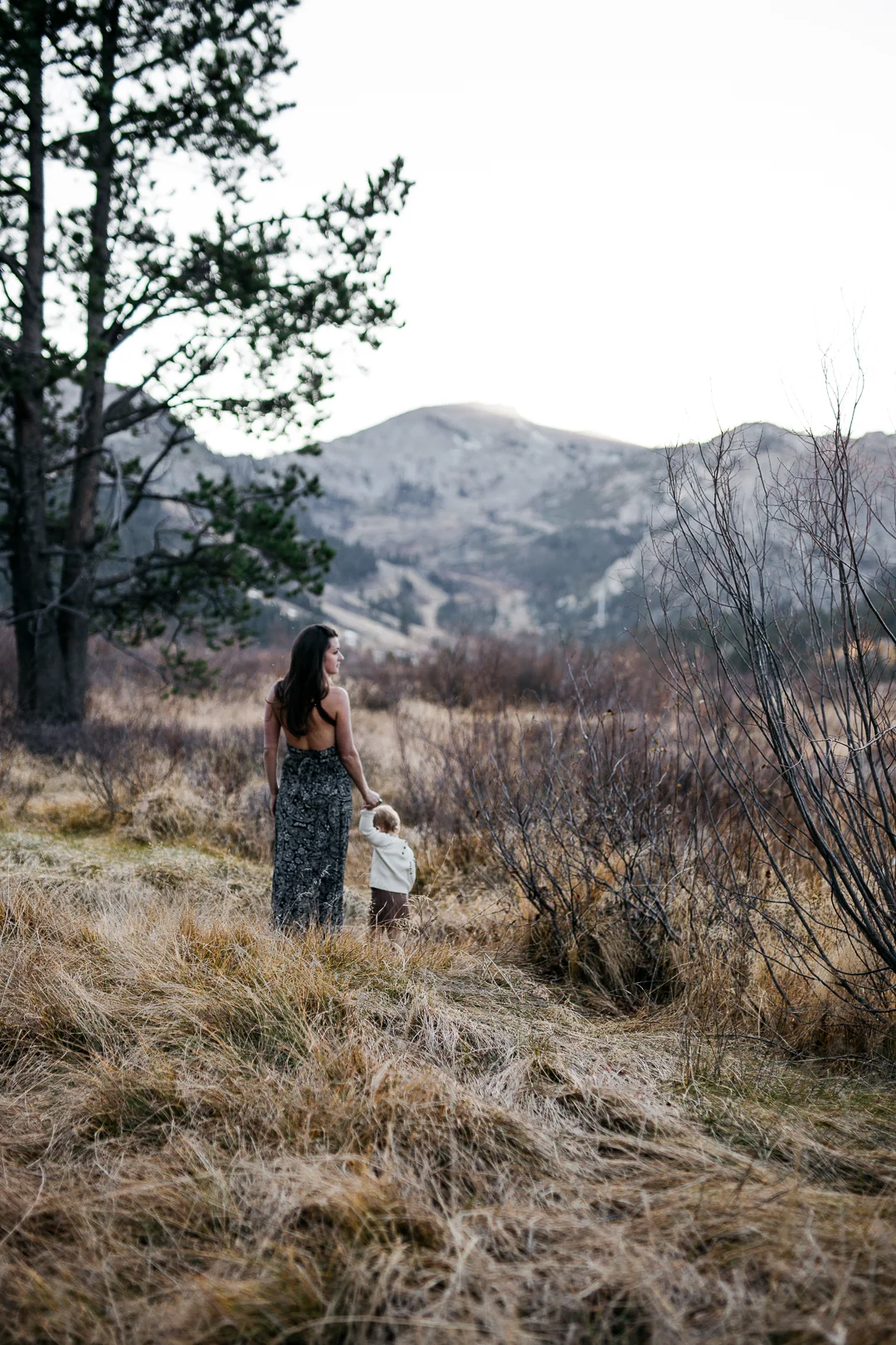 A woman and young child walking on a mountain trail surrounded by dry grass and bushes, with mountains in the background.