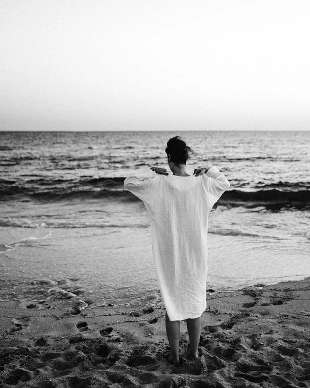 A woman in a white dress standing on the beach, facing the ocean with her arms raised and hands on her shoulders, during what appears to be sunset or sunrise.
