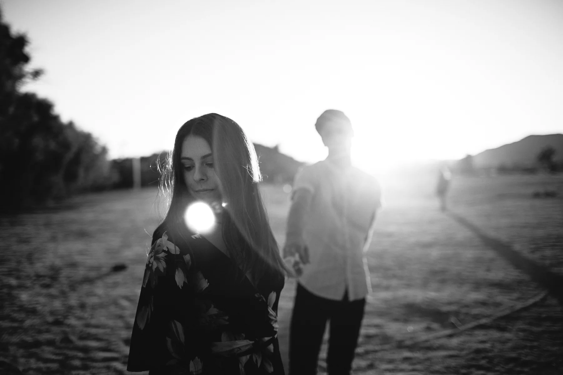 A young woman with long hair overlooking a landscape at sunset, with a young man in the background and the sun low on the horizon.